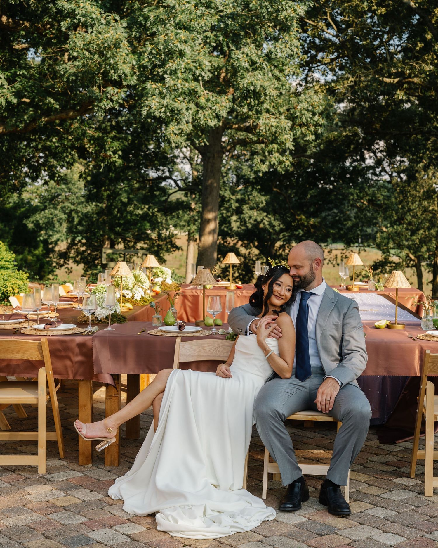A bride and groom sit together at an outdoor wedding reception table surrounded by lush trees and elegant terracotta decor.
