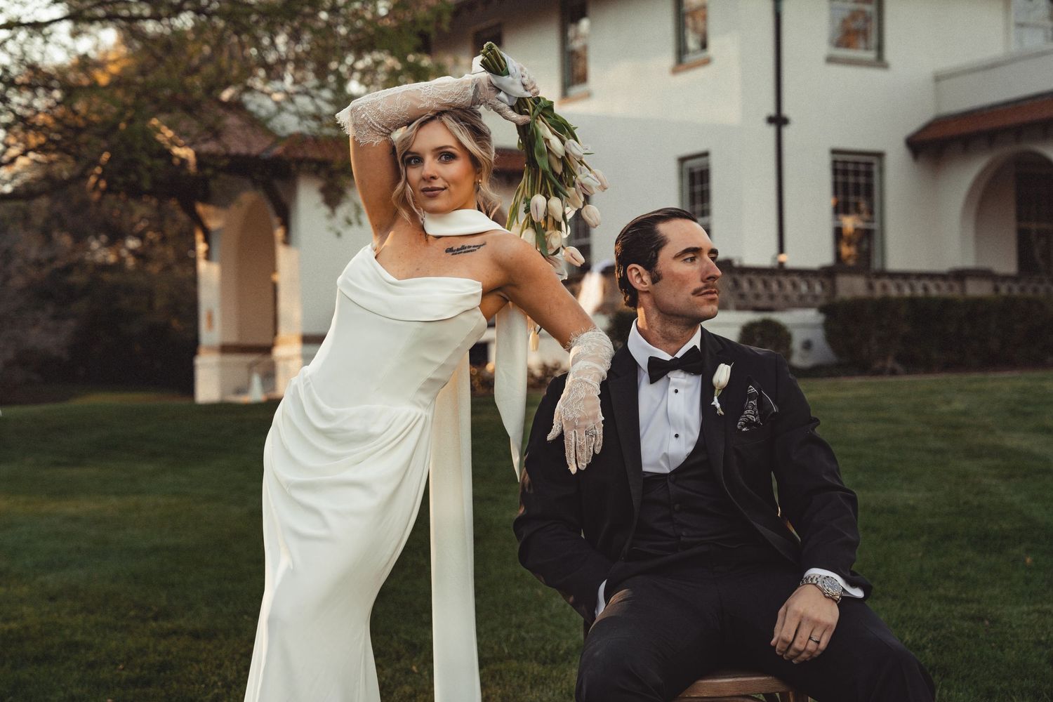 Bride in white jumpsuit and gloves poses with groom in black tuxedo on lush green lawn outside elegant estate.