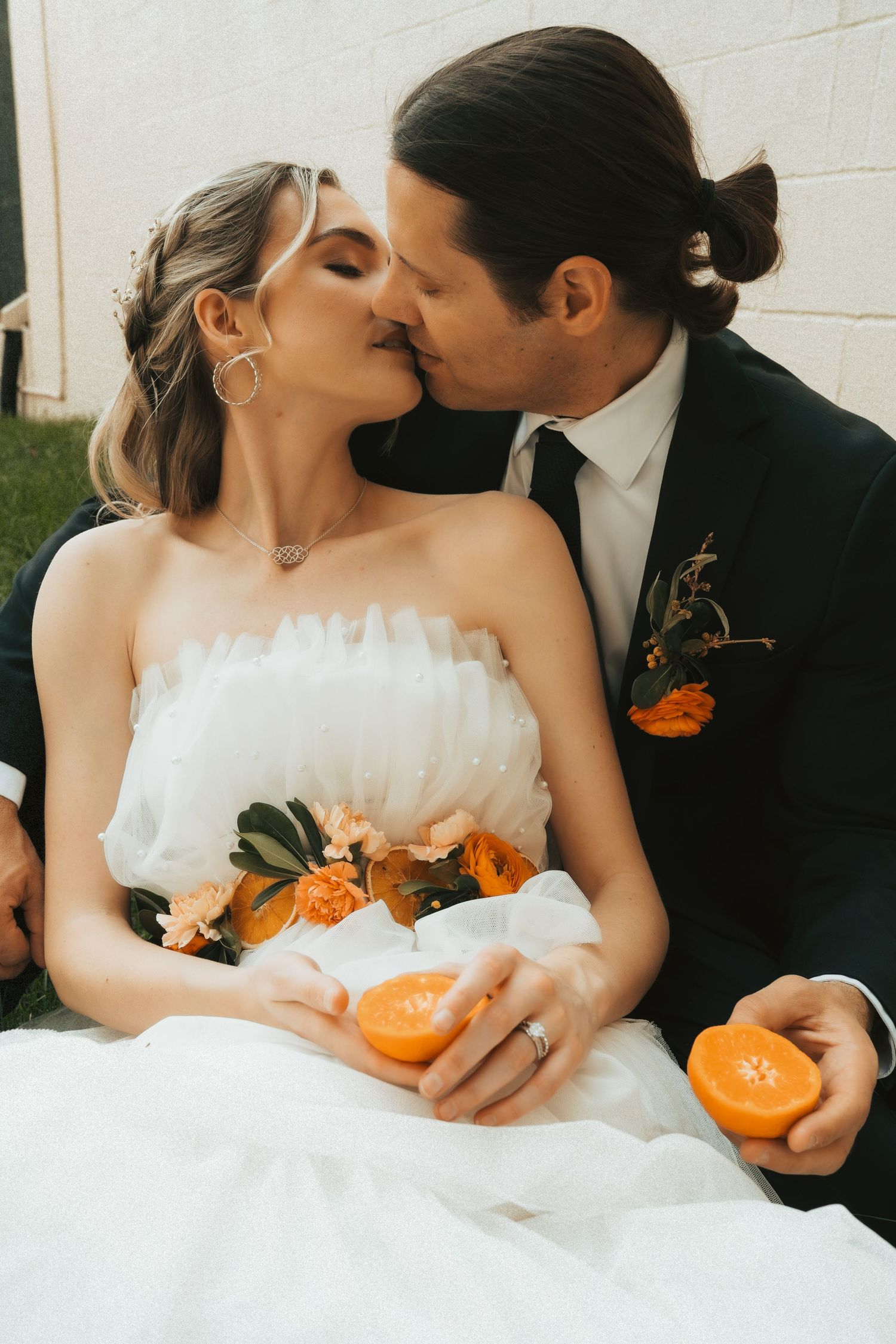 Wedding couple kissing outdoors, bride in white feather gown holding orange slices, groom in dark suit with orange boutonniere.