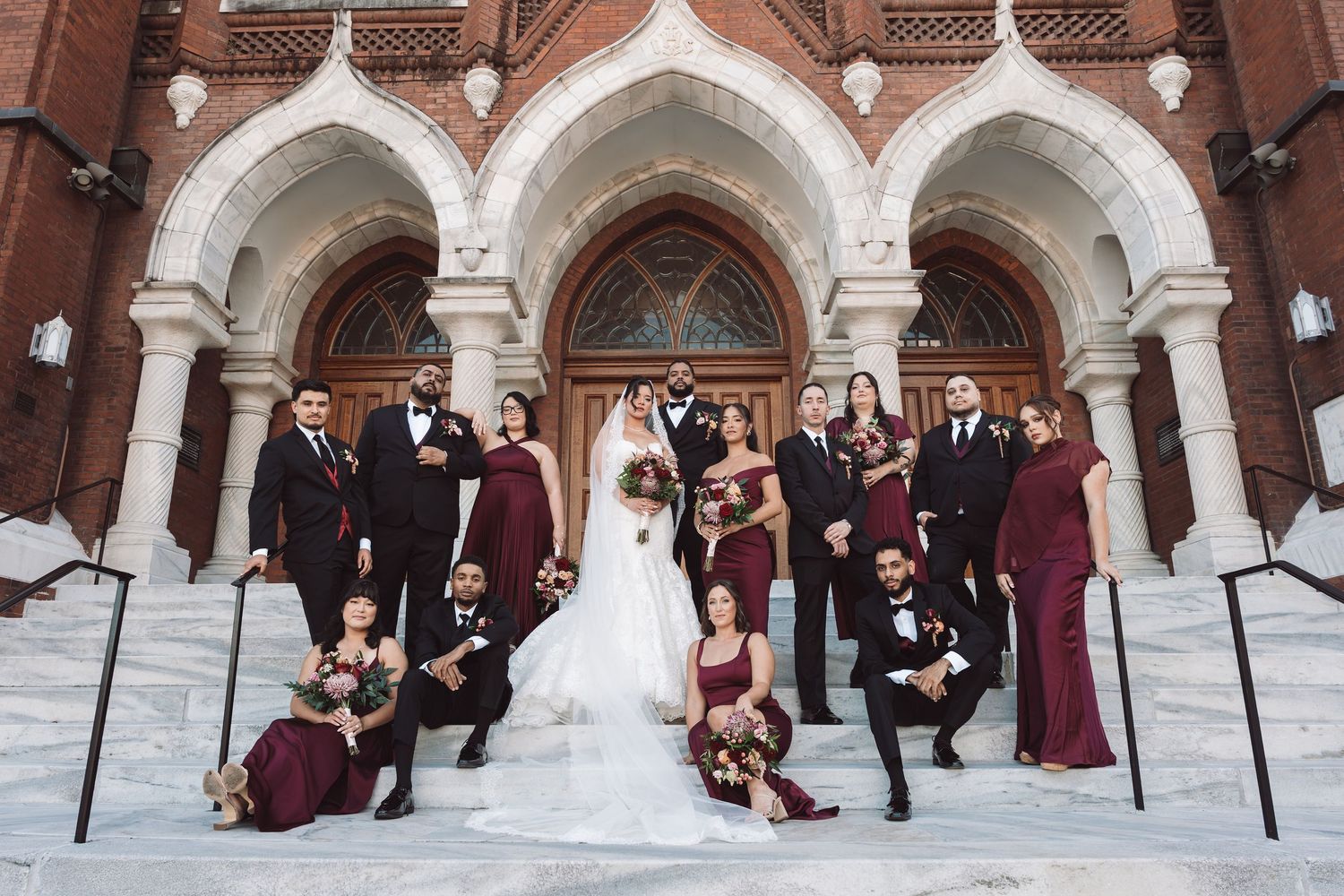 Wedding party in burgundy and black attire poses on church steps with bride and groom at center.