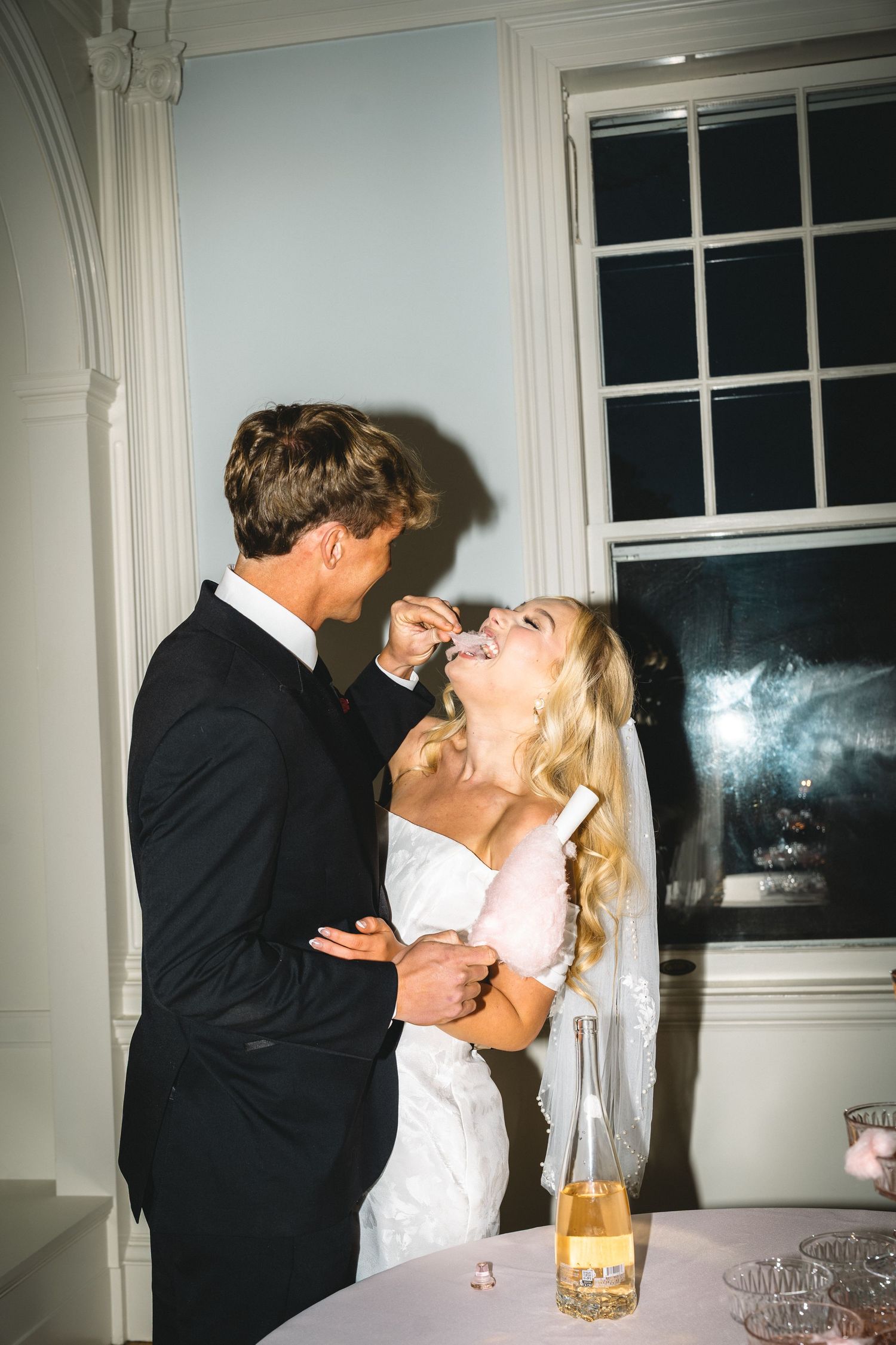 Bride and groom sharing wedding cake at reception, blonde bride in white gown laughing as groom feeds her.