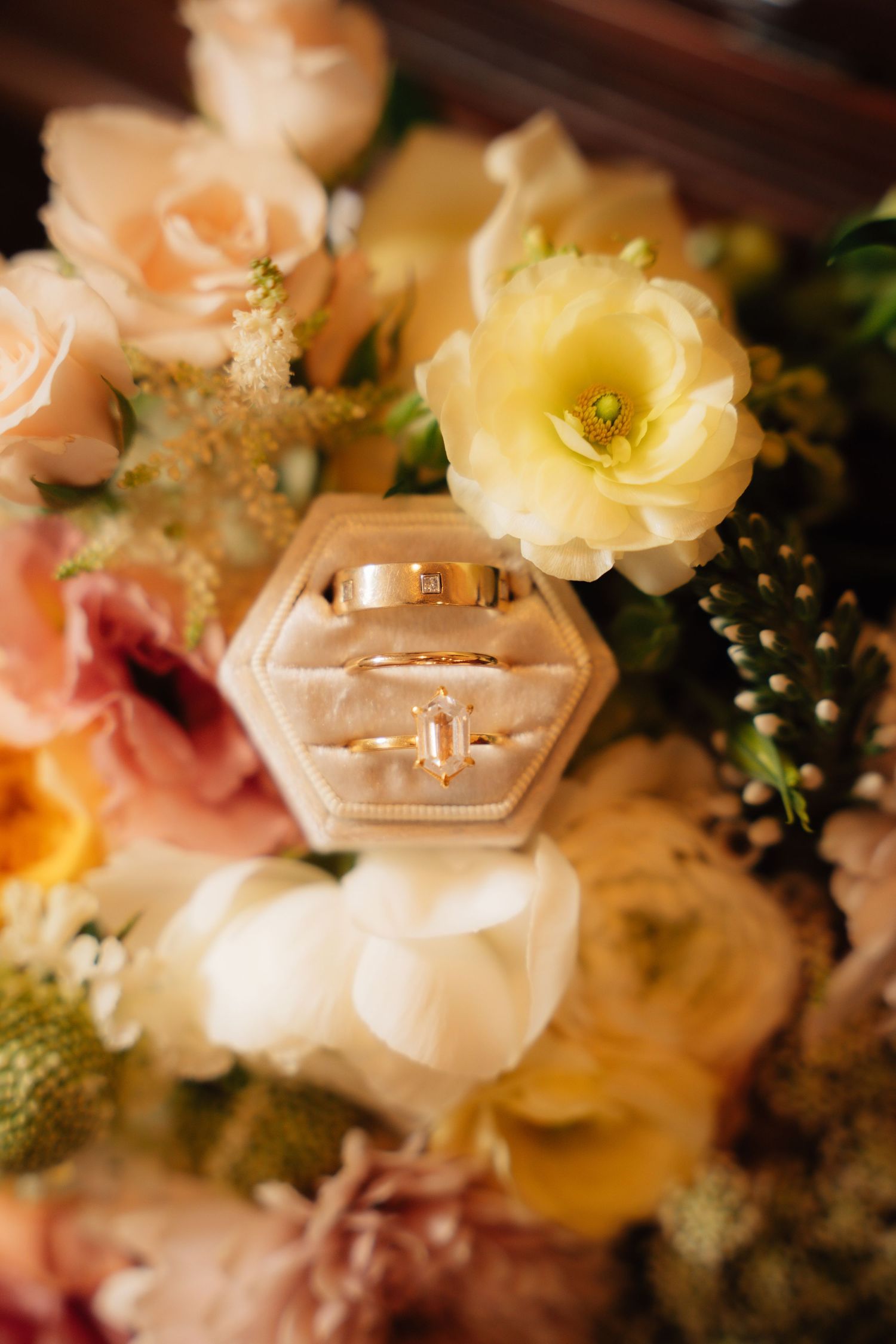 Gold wedding bands and diamond engagement ring in velvet box on colorful bridal bouquet with roses.