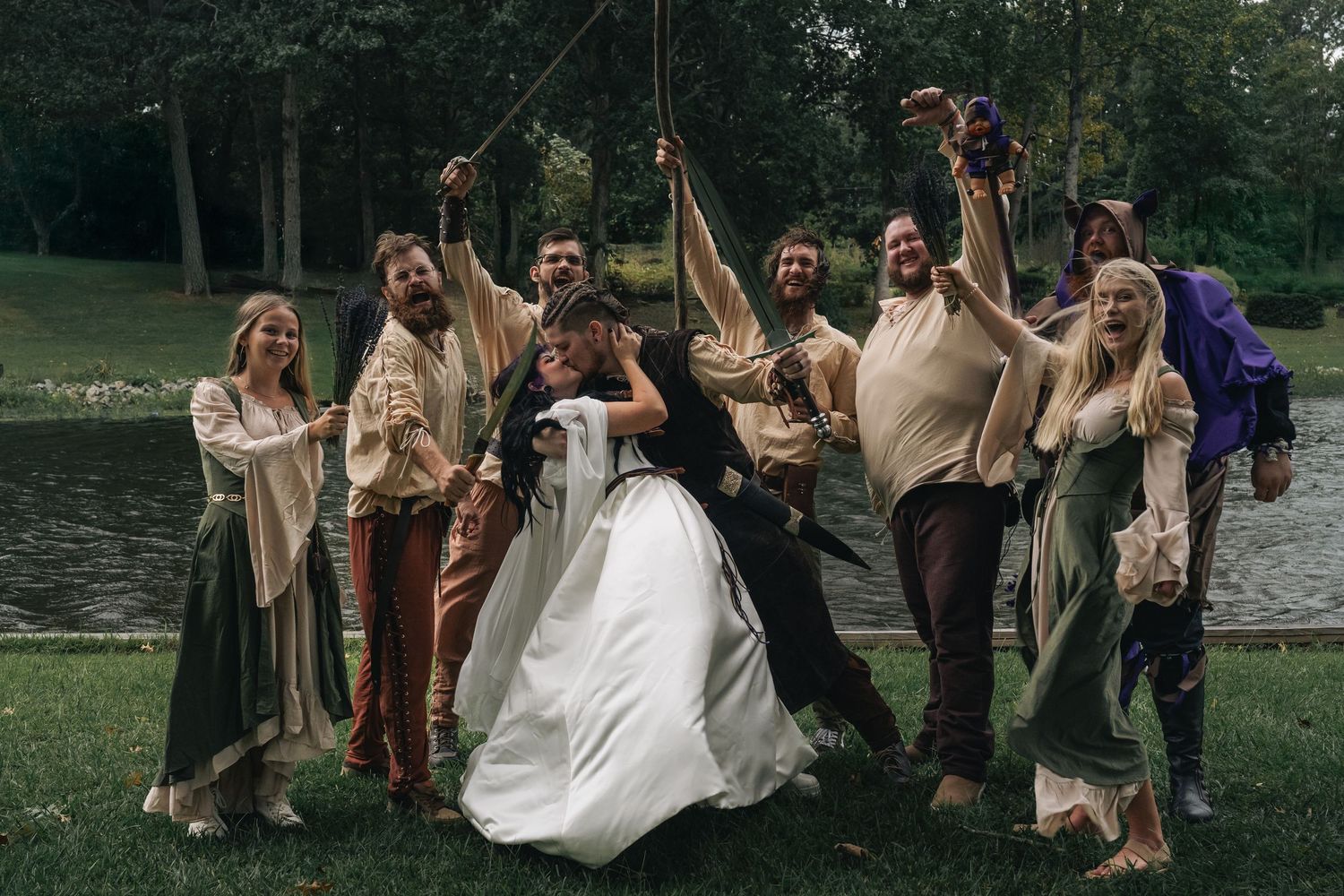 Medieval-themed wedding party celebrates outdoors near a pond, with guests in costume raising props around the bride and groom.