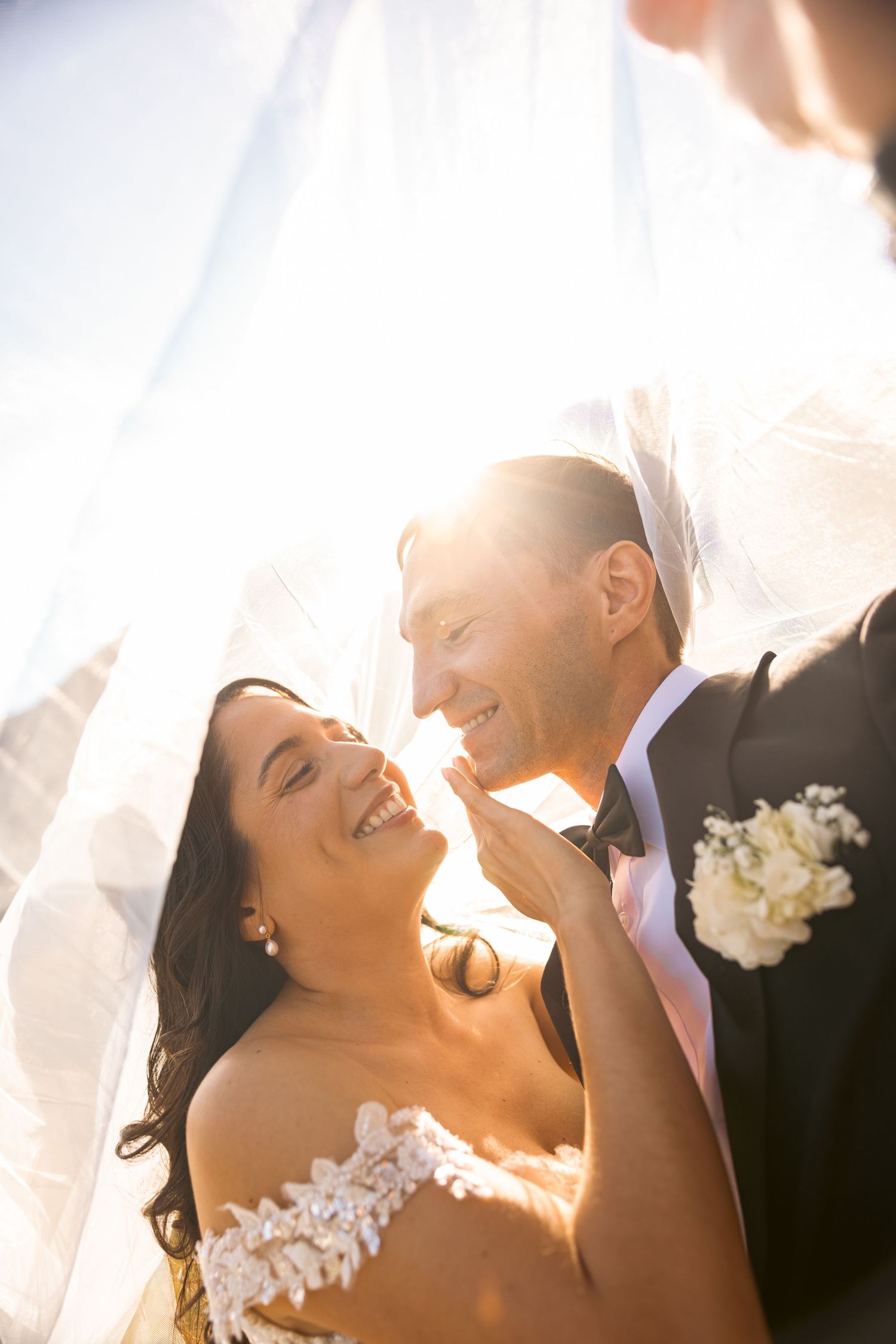 Bride and groom laughing joyfully under a veil with golden sunlight glowing behind them on their wedding day.