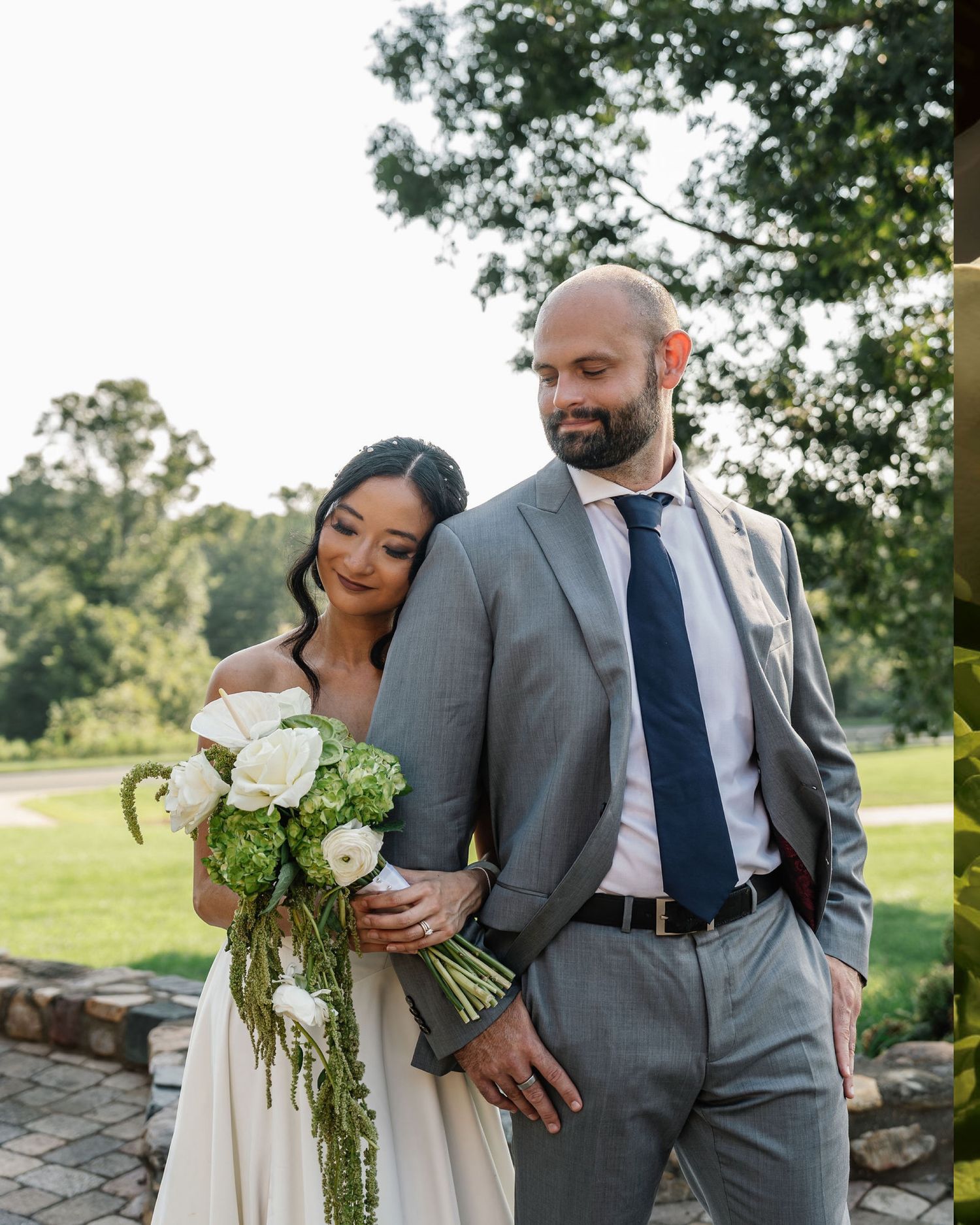 A bride and groom pose outdoors, she holds a white and green bouquet, he wears a gray suit with navy tie.