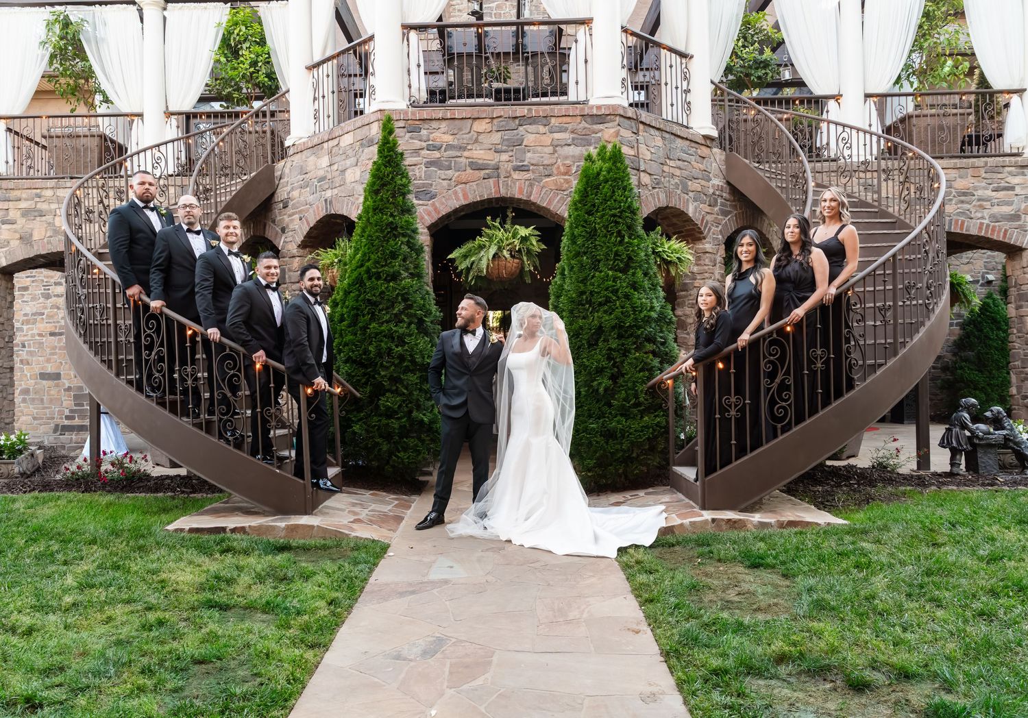 Bride and groom pose on stone path flanked by wedding party on twin spiral staircases at elegant venue.