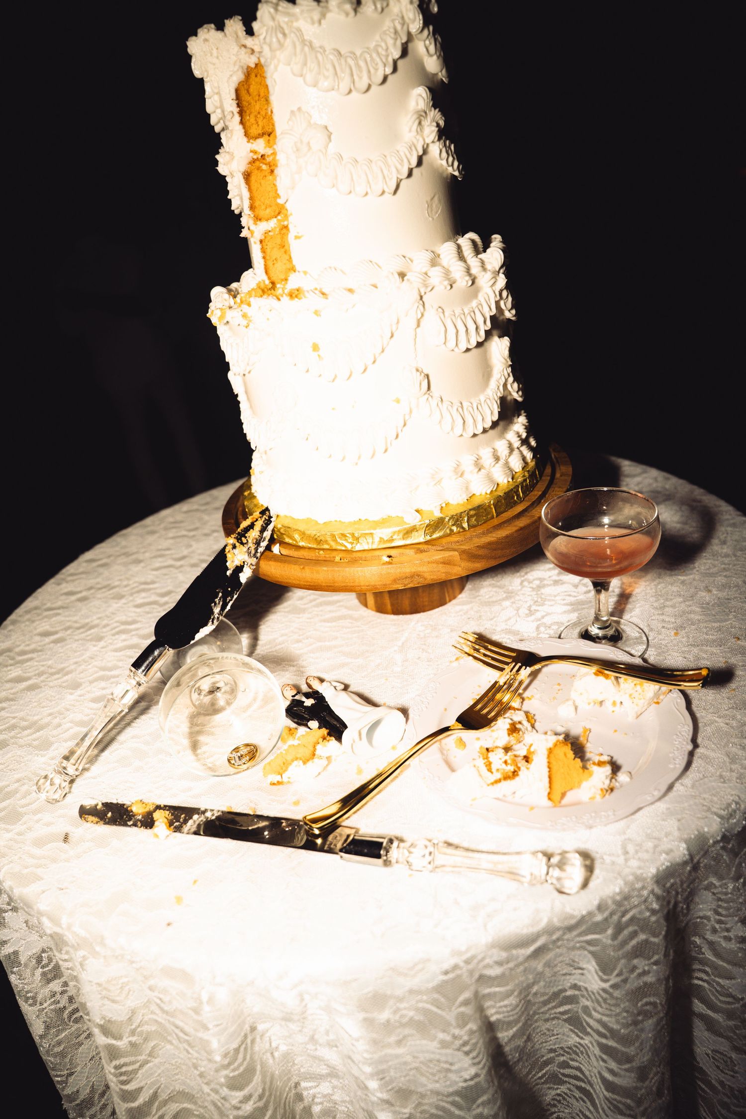Elegant white multi-tier wedding cake with gold accents on lace tablecloth, with cake server, forks, and pink cocktail.