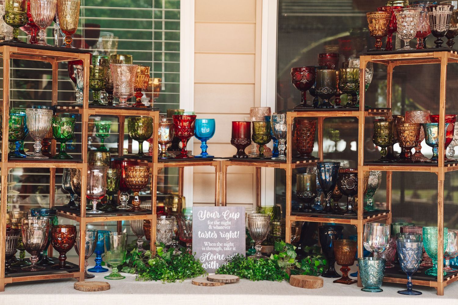 Colorful vintage glassware displayed on wooden shelves at an outdoor event, featuring goblets and cups in red, blue, and gold.