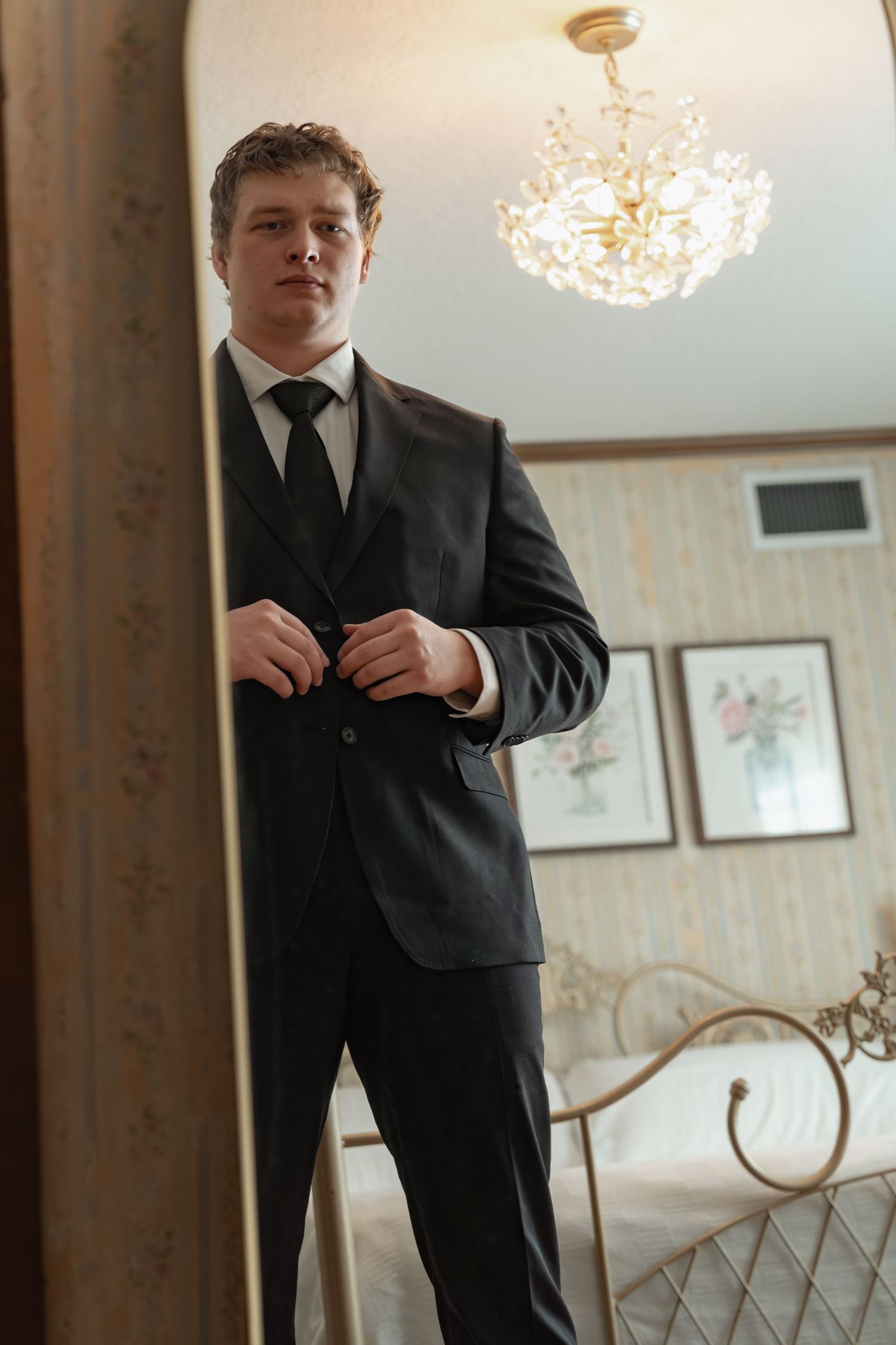 Young man in dark suit buttoning jacket while standing in elegant bedroom with chandelier.