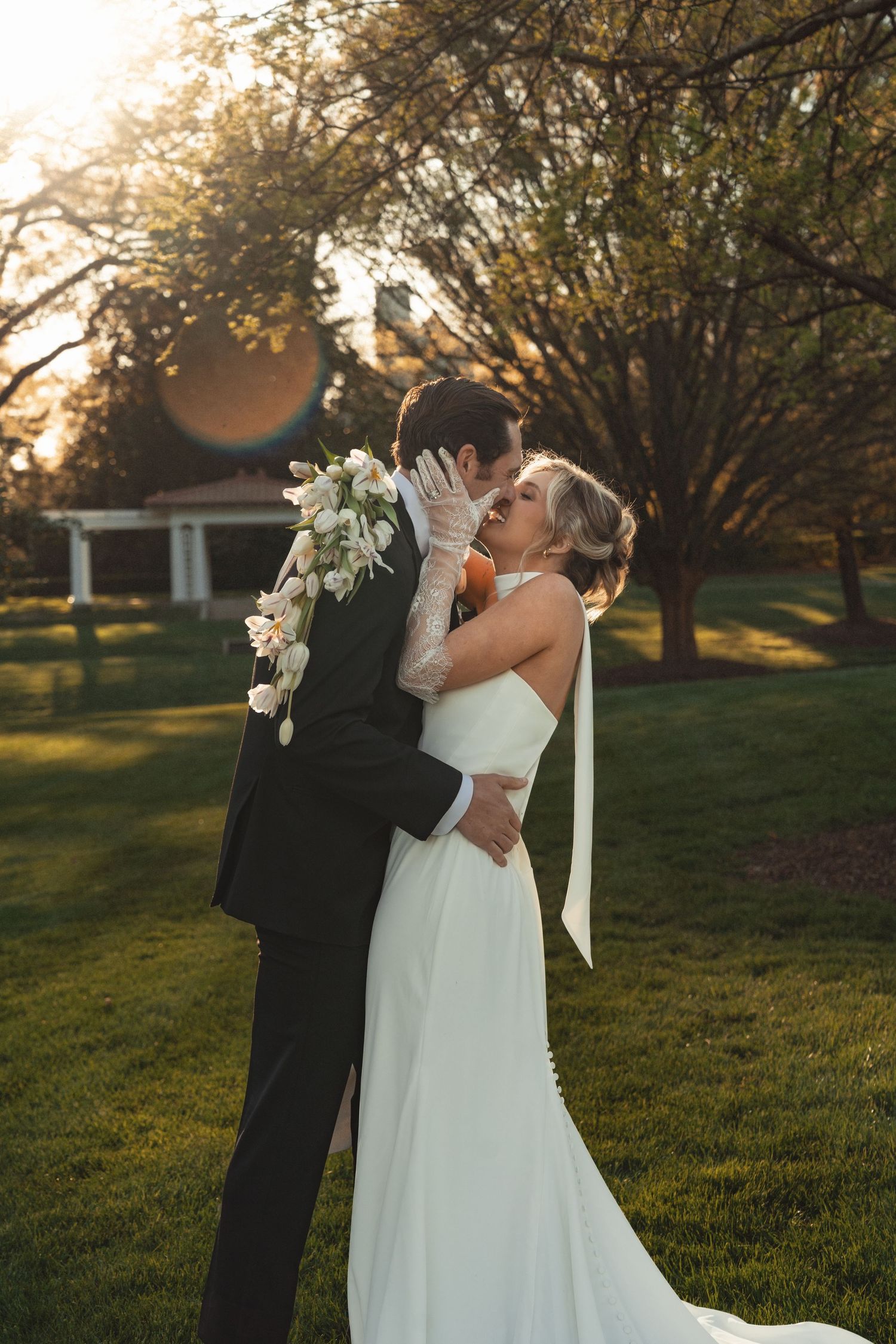 Bride and groom share a romantic kiss at golden hour on lush green lawn, bride wearing white gown and floral bouquet.