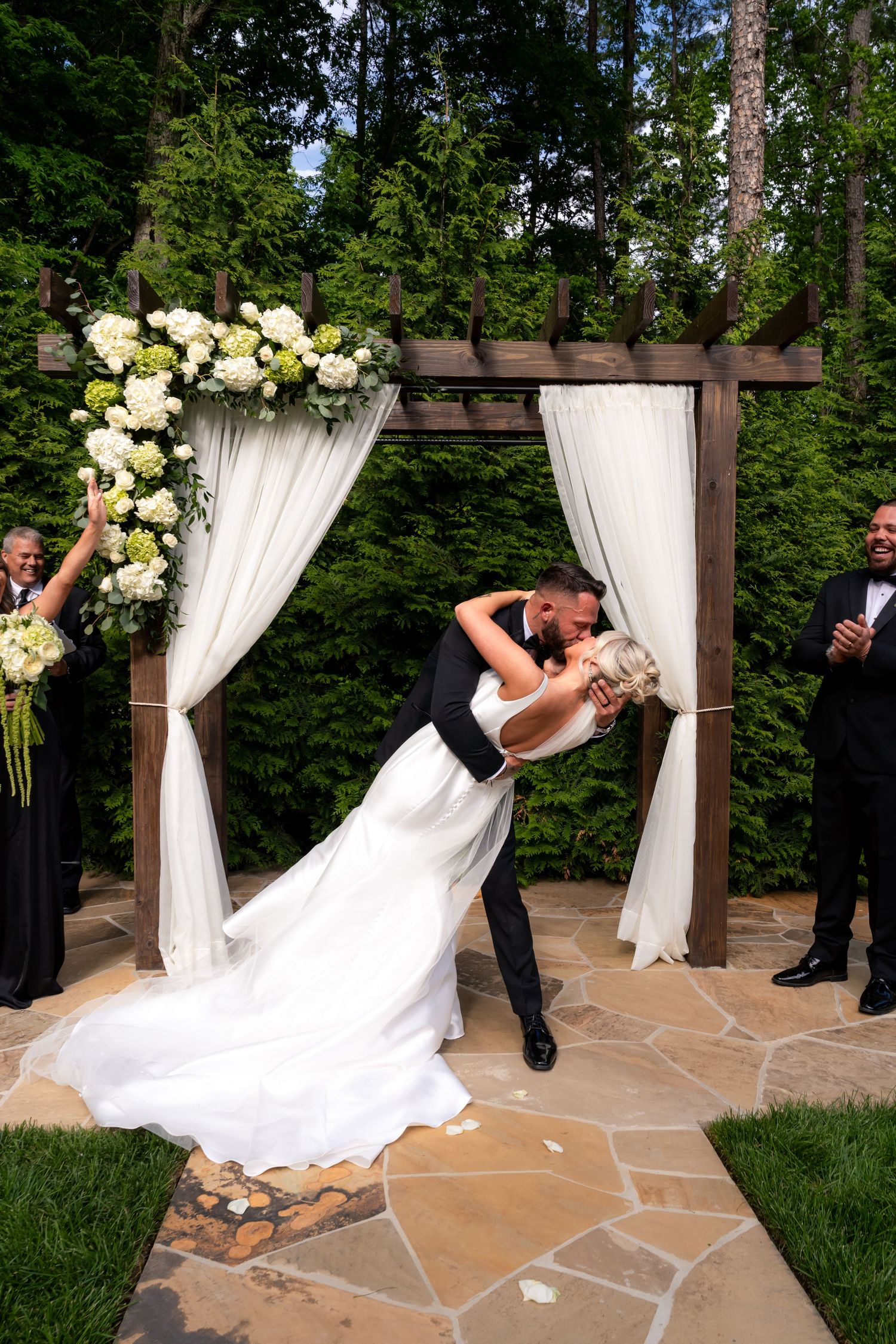 Groom dips bride for romantic kiss under floral arch with white drapes during outdoor wedding ceremony.