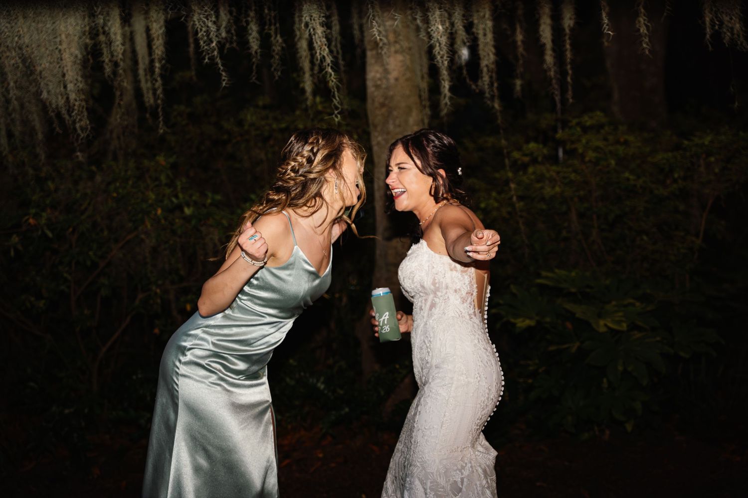 Bride in lace gown laughing with bridesmaid in sage satin dress outdoors at night wedding reception.