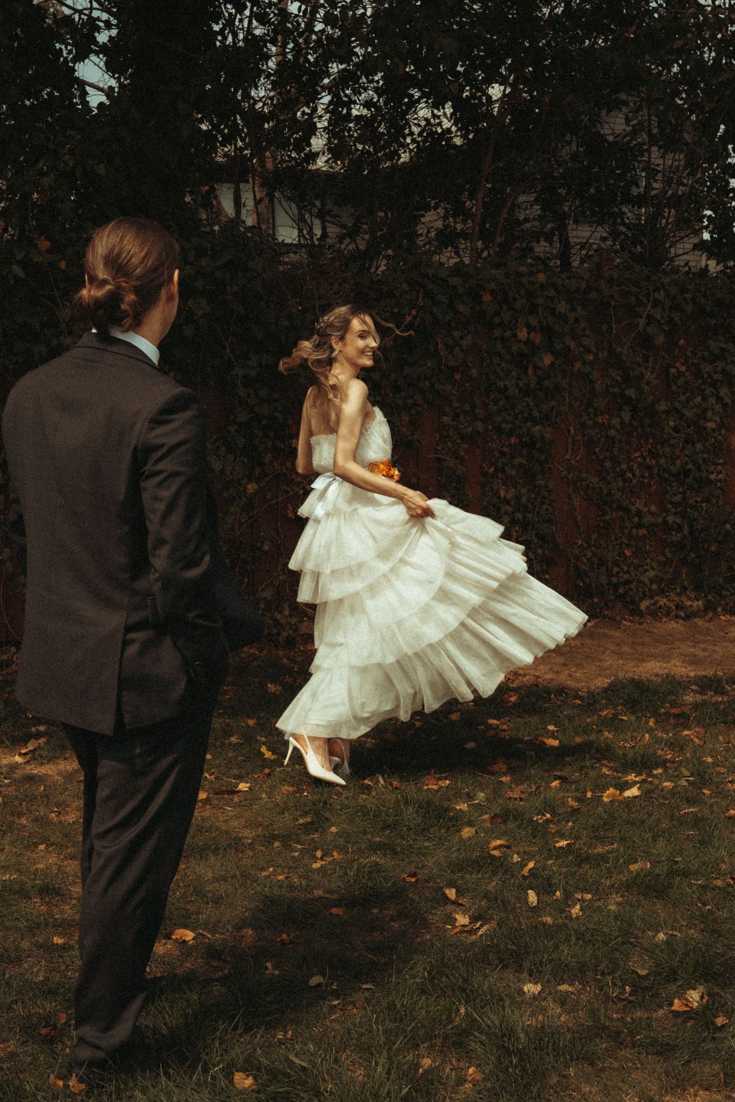 Bride in layered white tulle gown twirls joyfully in autumn garden as groom watches adoringly.