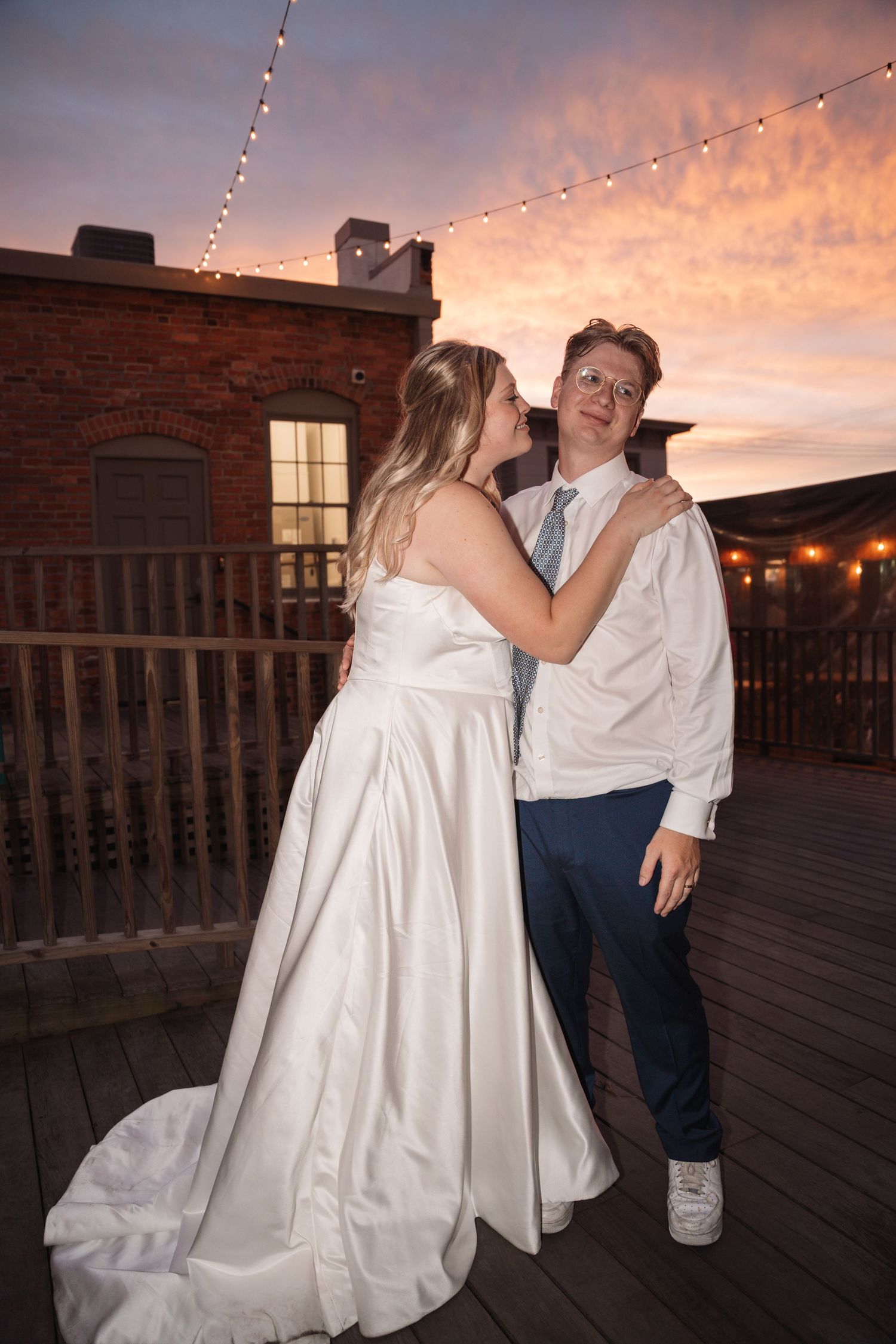 Bride in white gown kisses groom on cheek at sunset on rooftop deck with string lights and brick building backdrop.