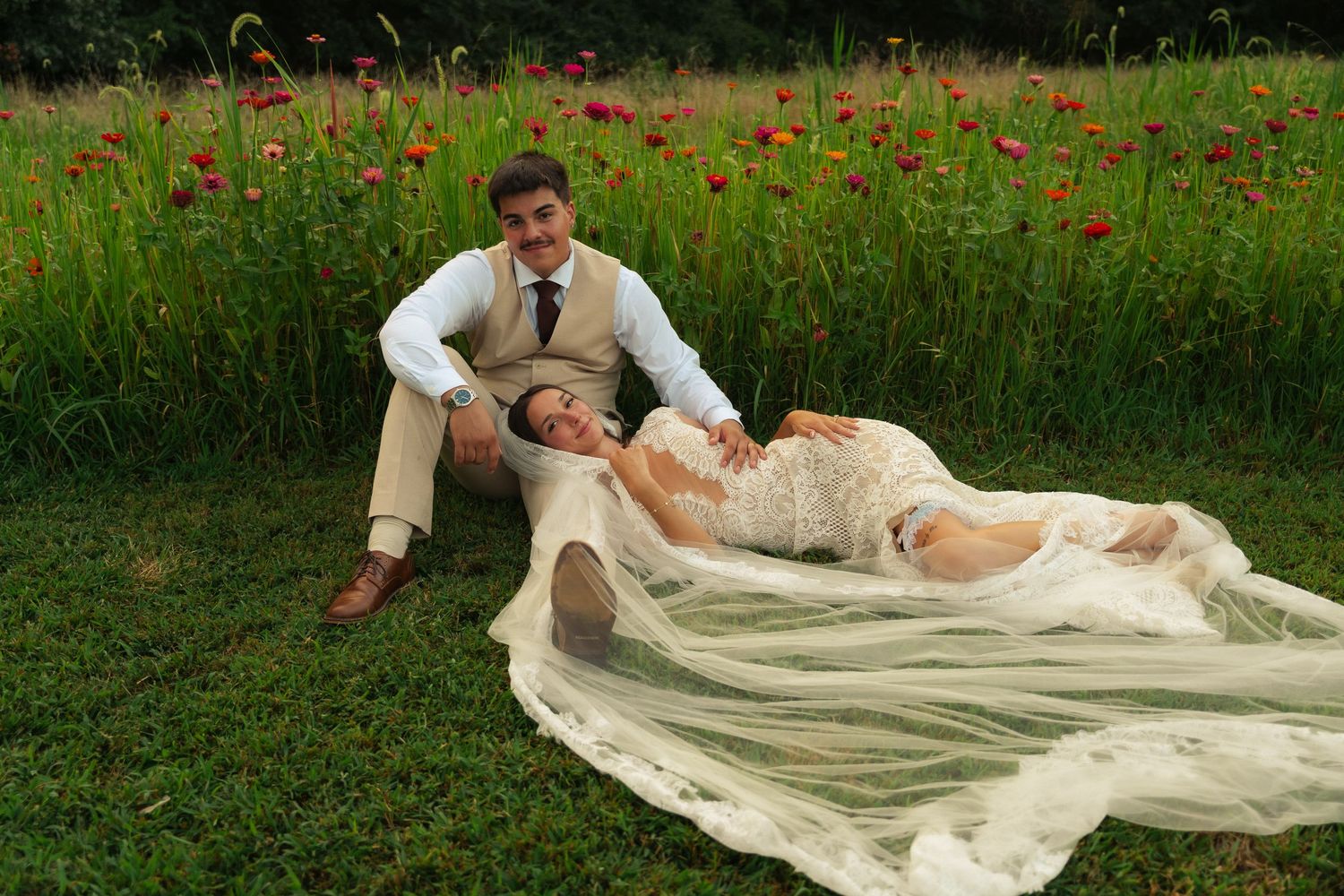 Bride in lace gown reclines on groom's lap in a lush green field with red poppies blooming behind them.