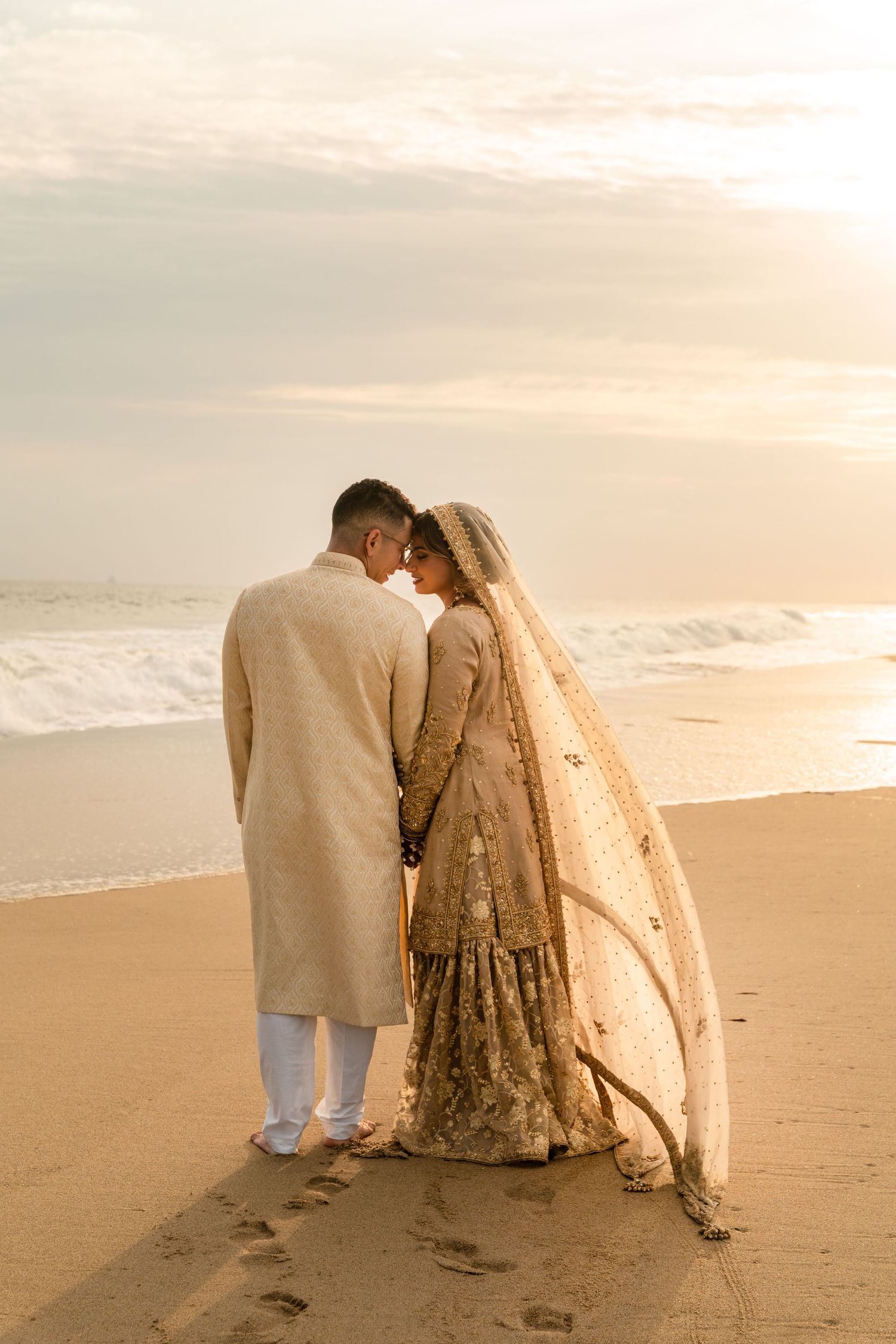Indian bride and groom in traditional gold attire hold hands on a sandy beach at sunset with ocean waves behind them.