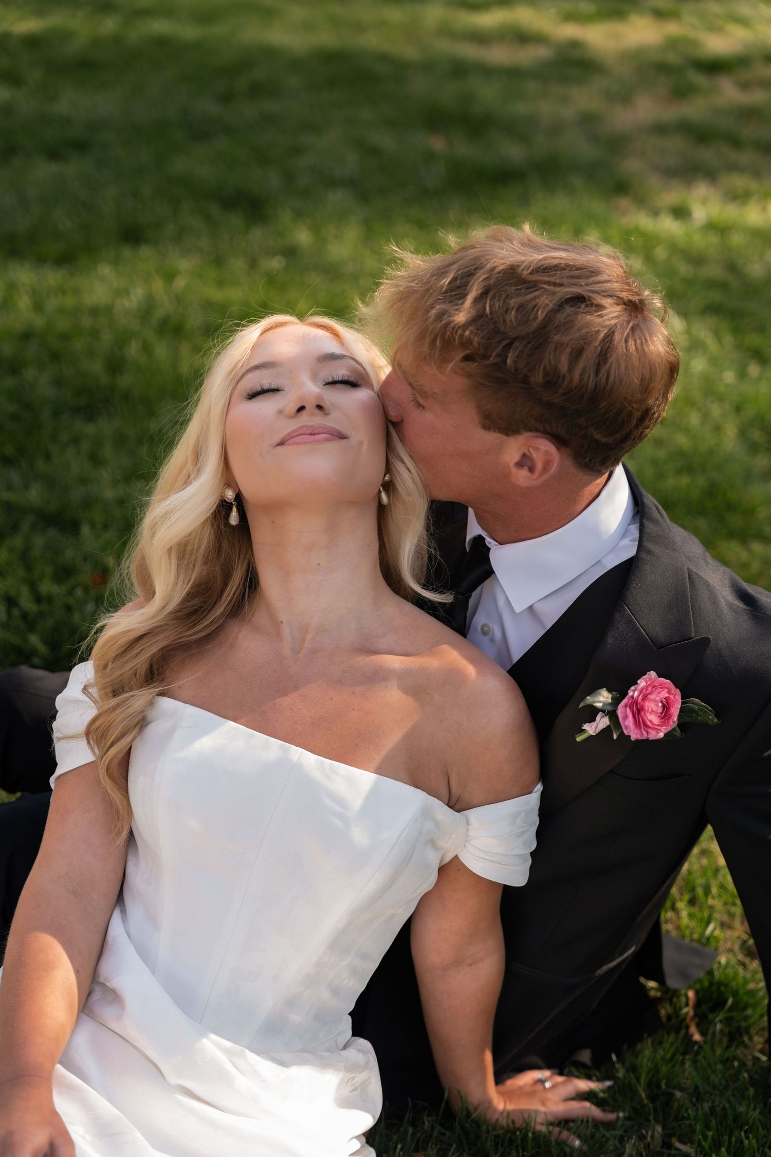 A groom in a black suit kisses his bride on the cheek as she smiles, sitting on green grass outdoors.