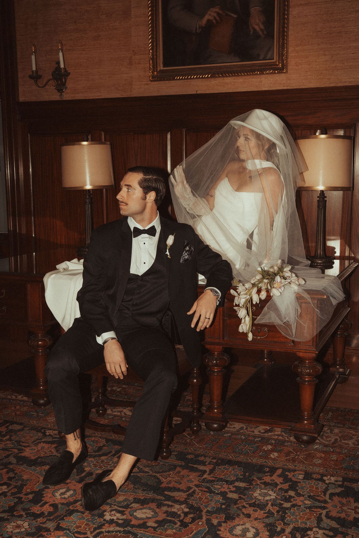 Bride in white gown and veil sits beside groom in black tuxedo in an elegant wood-paneled room with vintage decor.