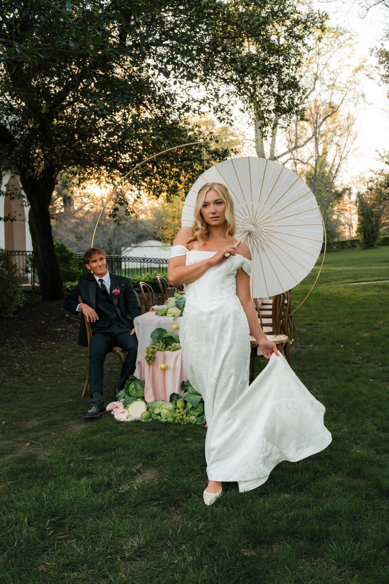 Bride in white off-shoulder gown holds parasol while groom sits in chair during golden hour outdoor wedding.