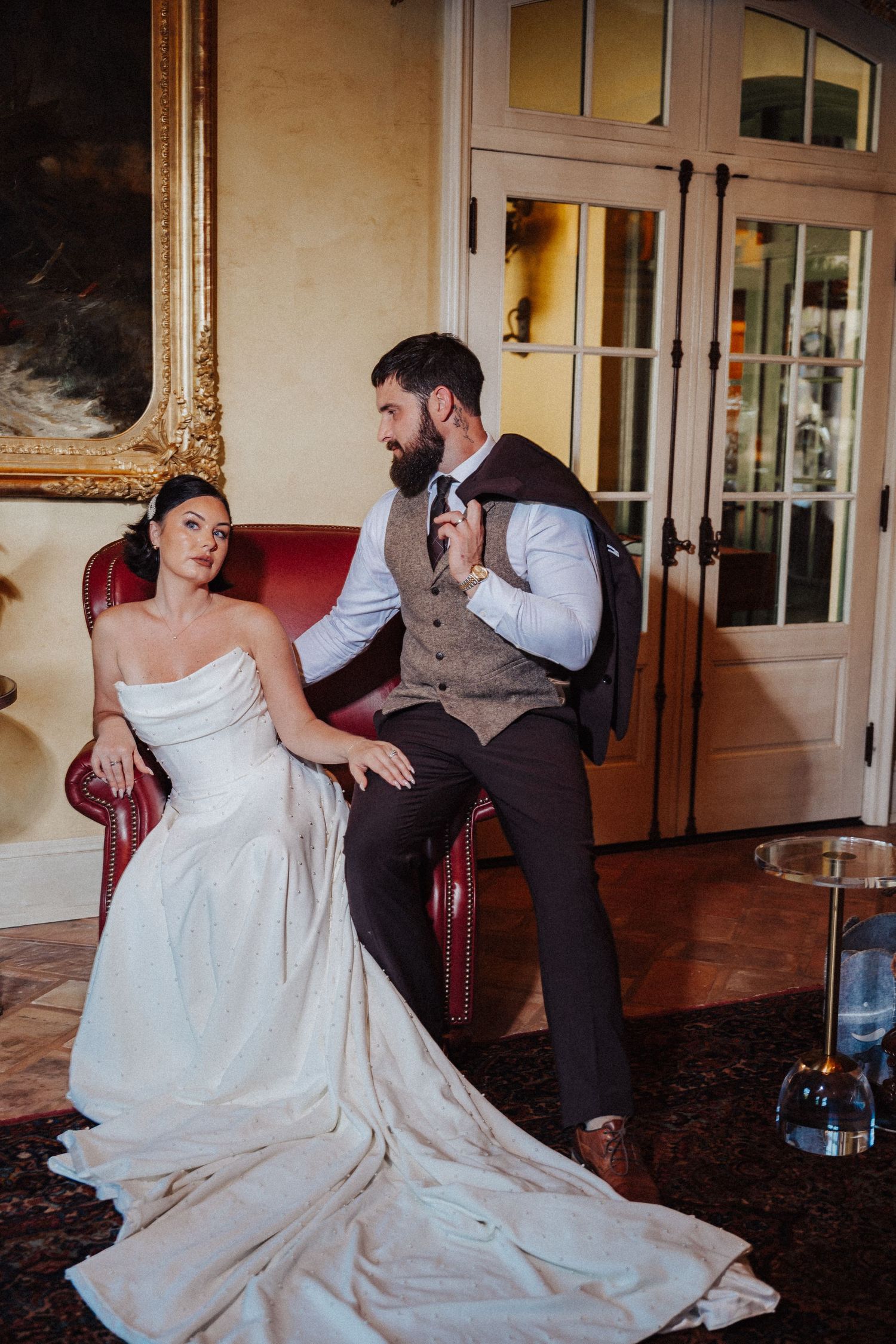 Bride in strapless white gown sits beside bearded groom in vest on red leather chair in elegant room.