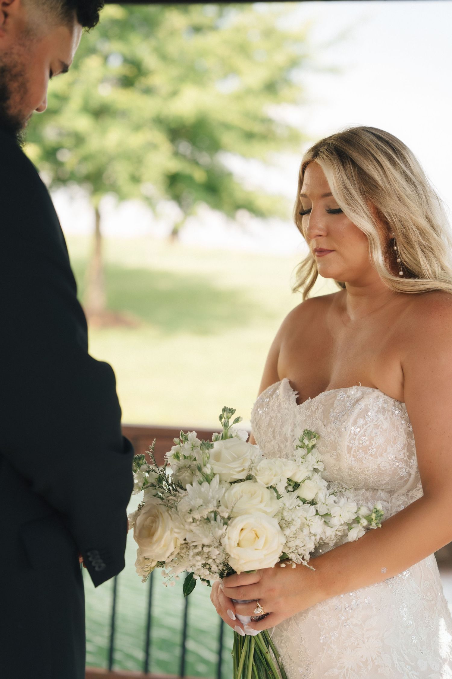 Blonde bride in strapless beaded gown holds white rose bouquet outdoors.