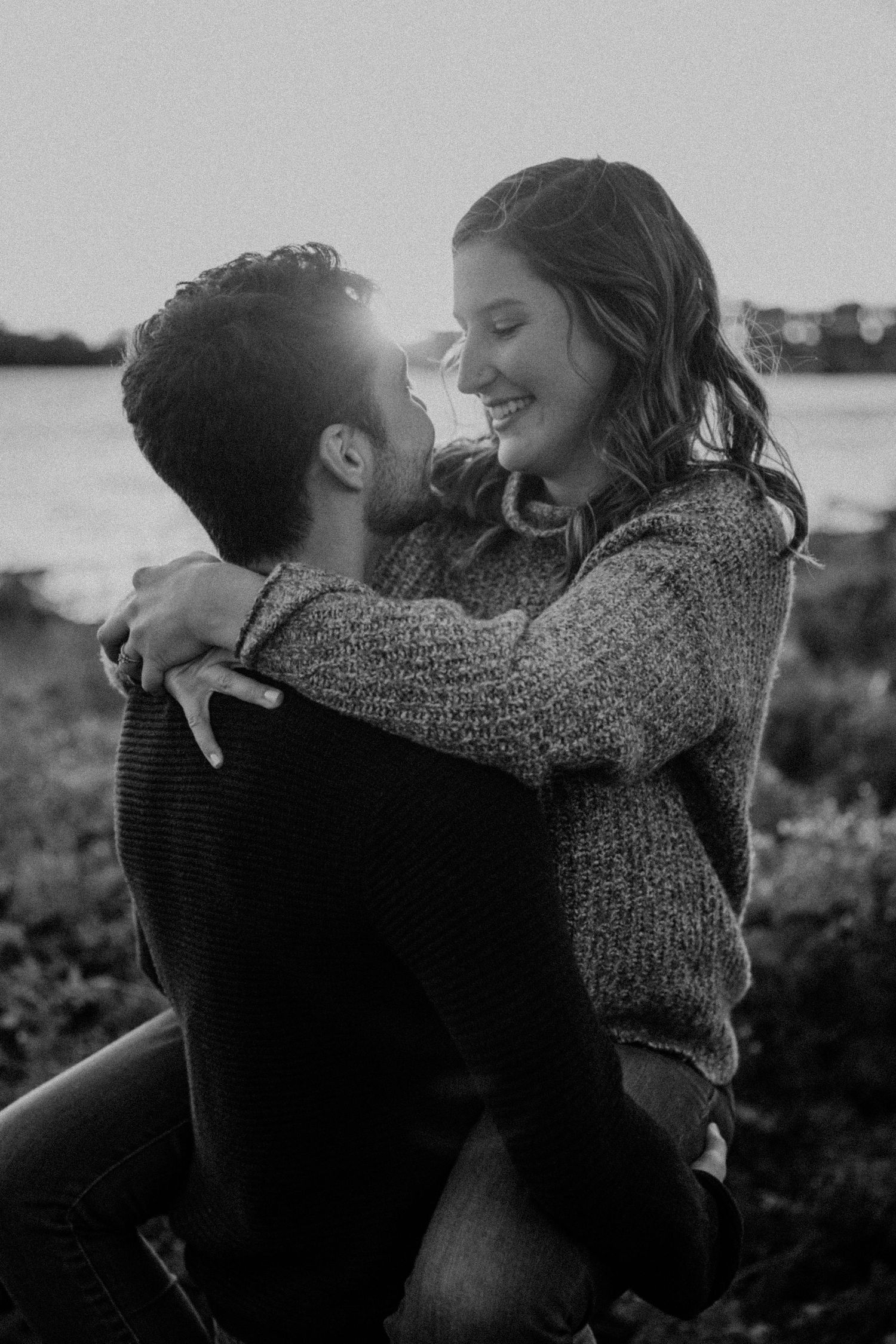 Black and white photo of embracing couple laughing together by waterfront.