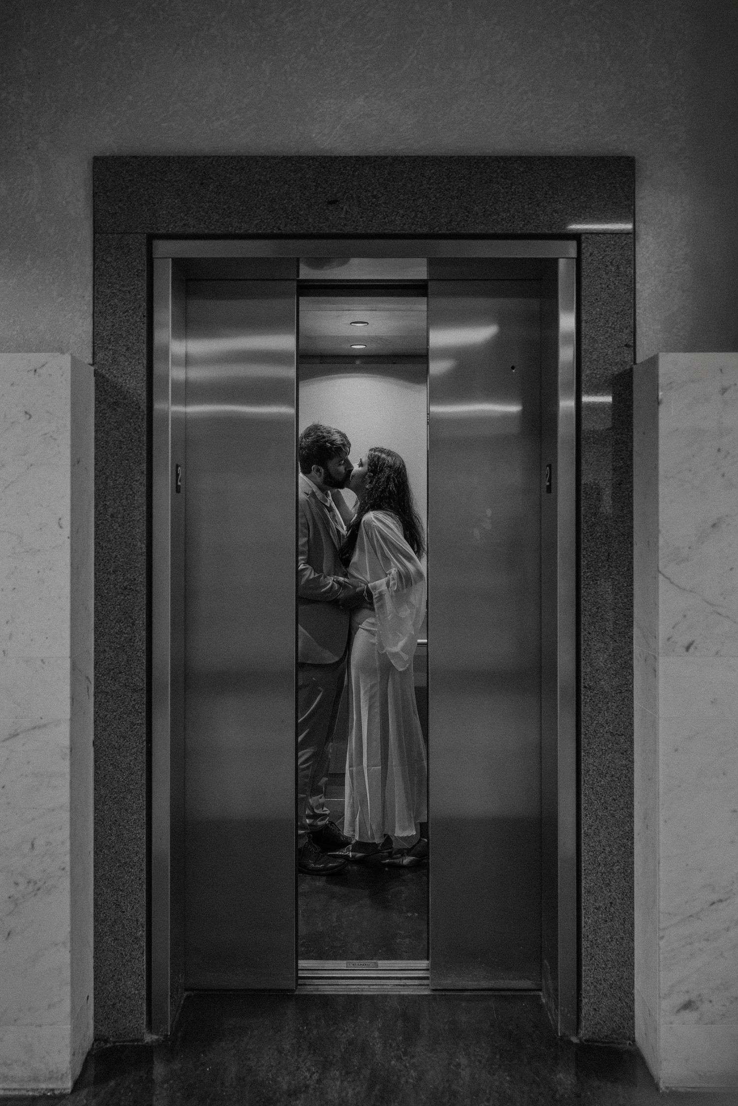Black and white photo of a couple kissing inside an elevator with open doors.