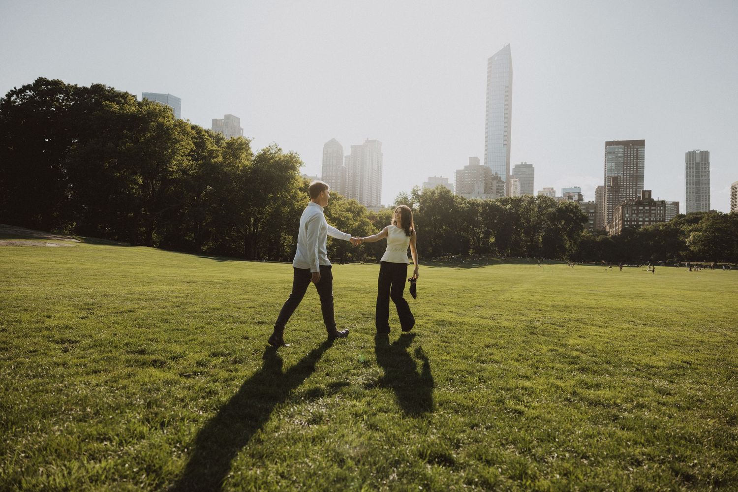 Couple holding hands walking on green grass in Central Park with NYC skyline in the background.