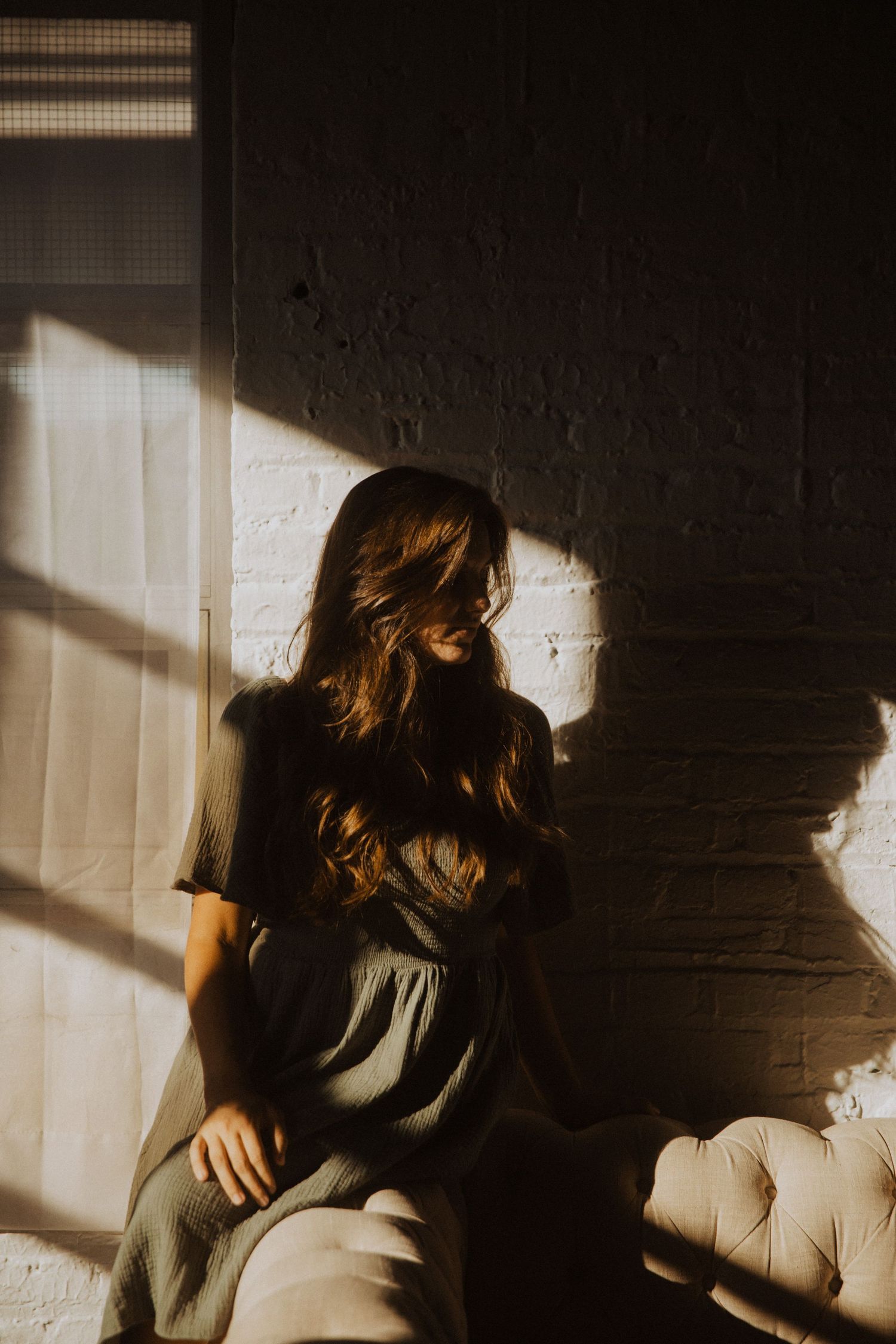Woman with long wavy hair in gray dress sits in dramatic sunlight against white brick wall.