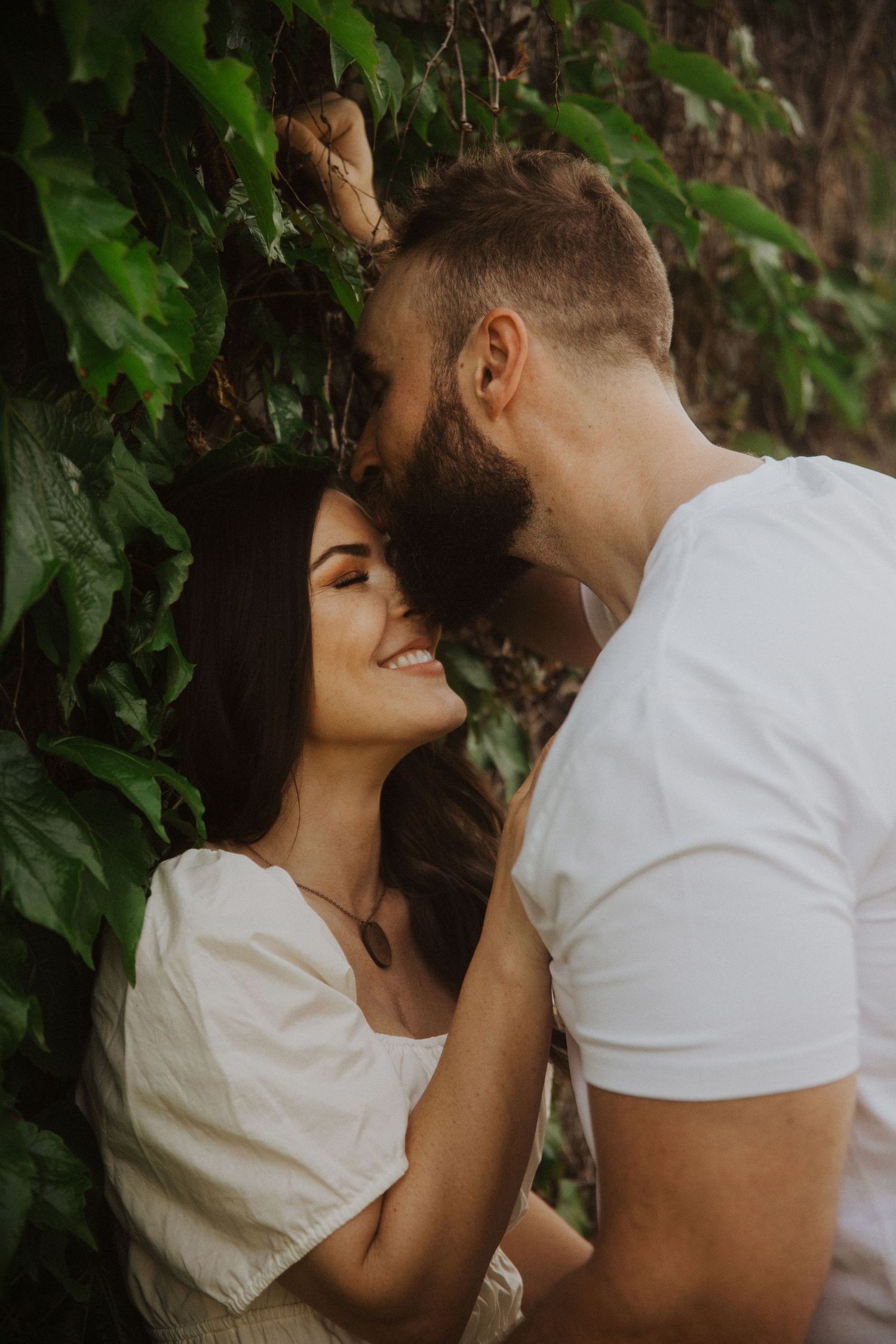 Romantic couple in white clothing embracing near a wall covered in lush green vines.