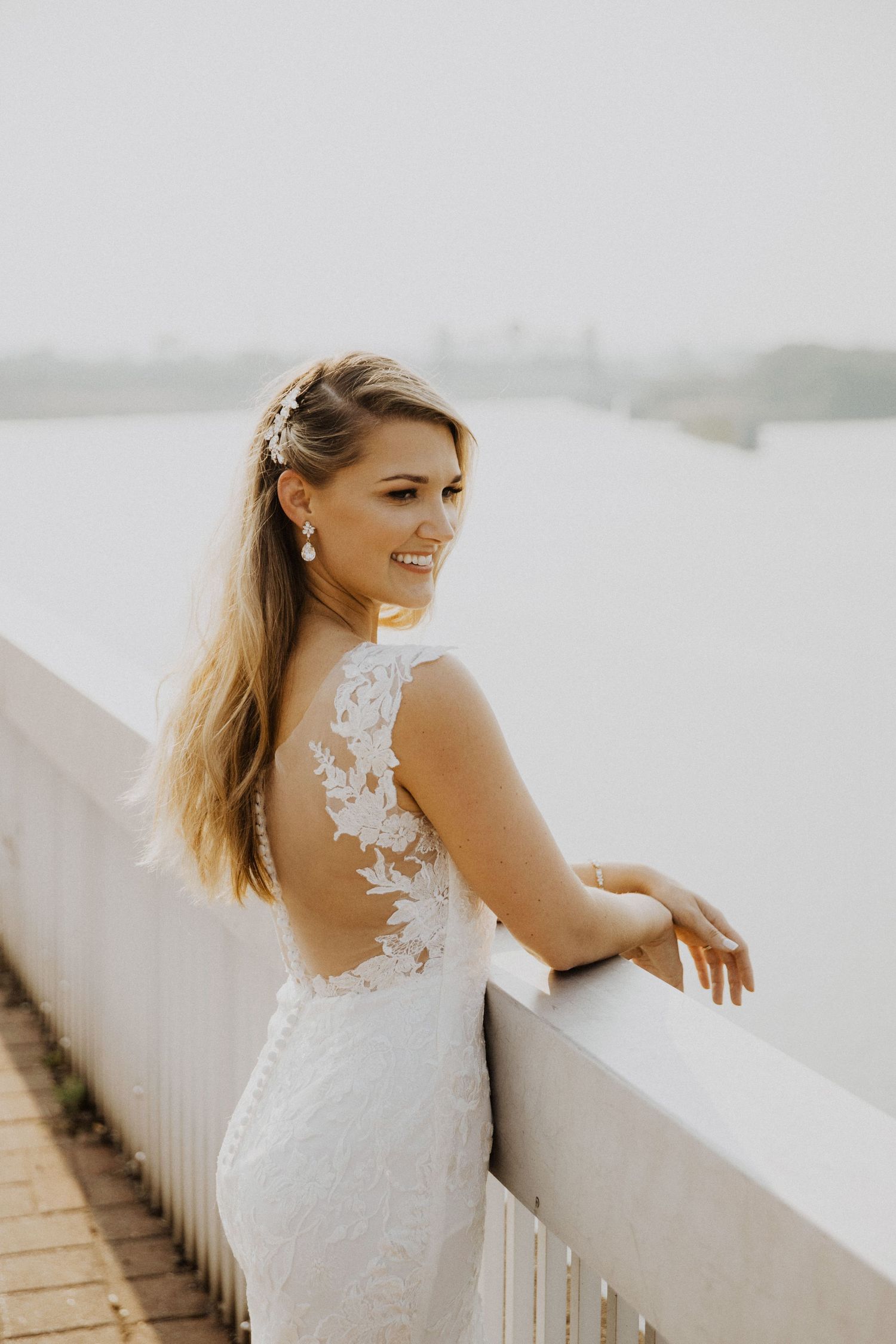 Bride in lace dress poses against white railing during sunset.