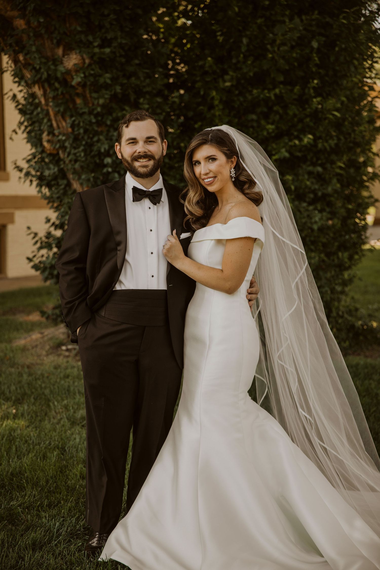 Elegant bride in white gown and veil poses with groom in tuxedo against garden backdrop.