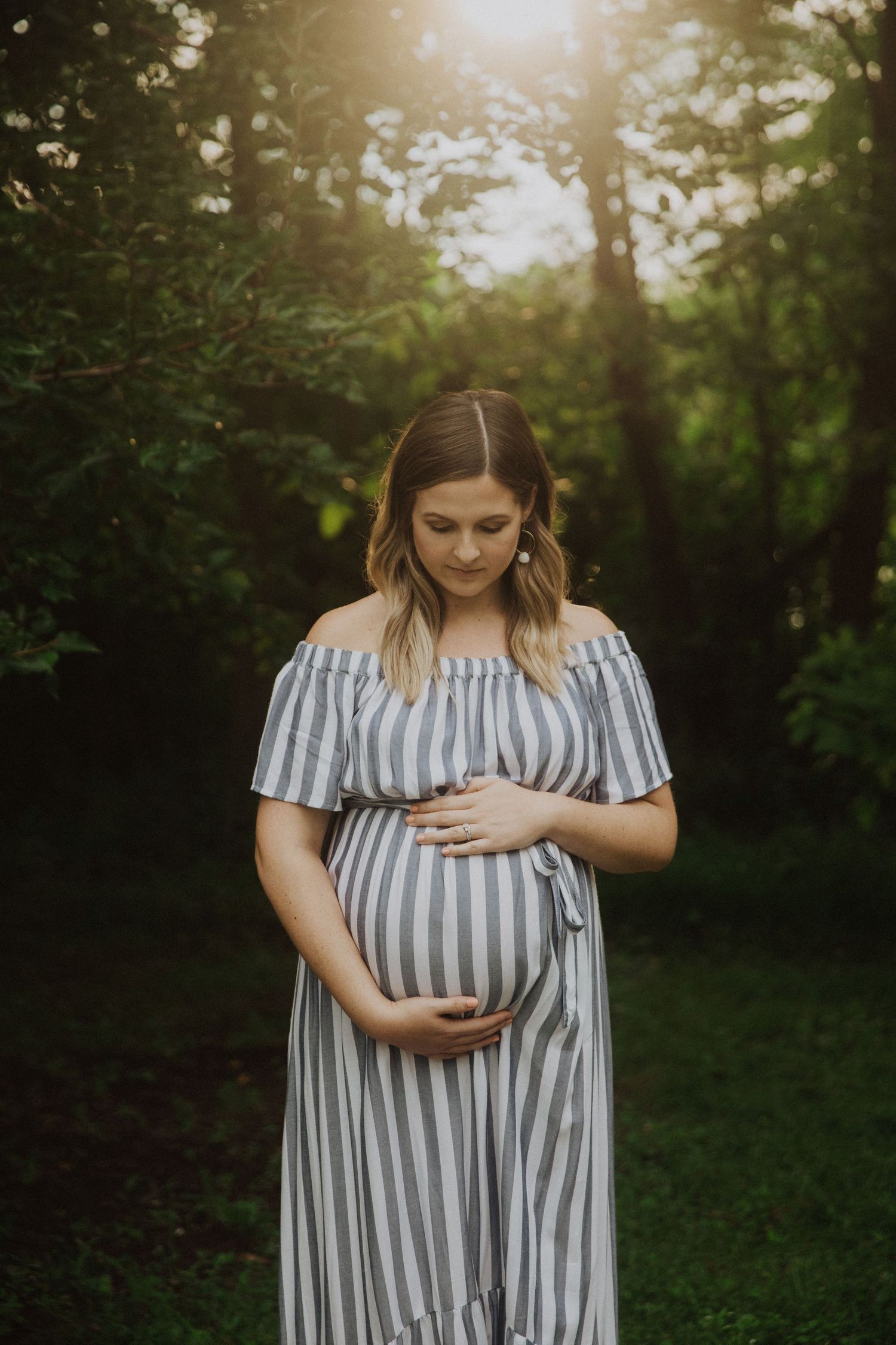 Pregnant woman in striped off-shoulder dress cradling baby bump outdoors in golden sunlight.