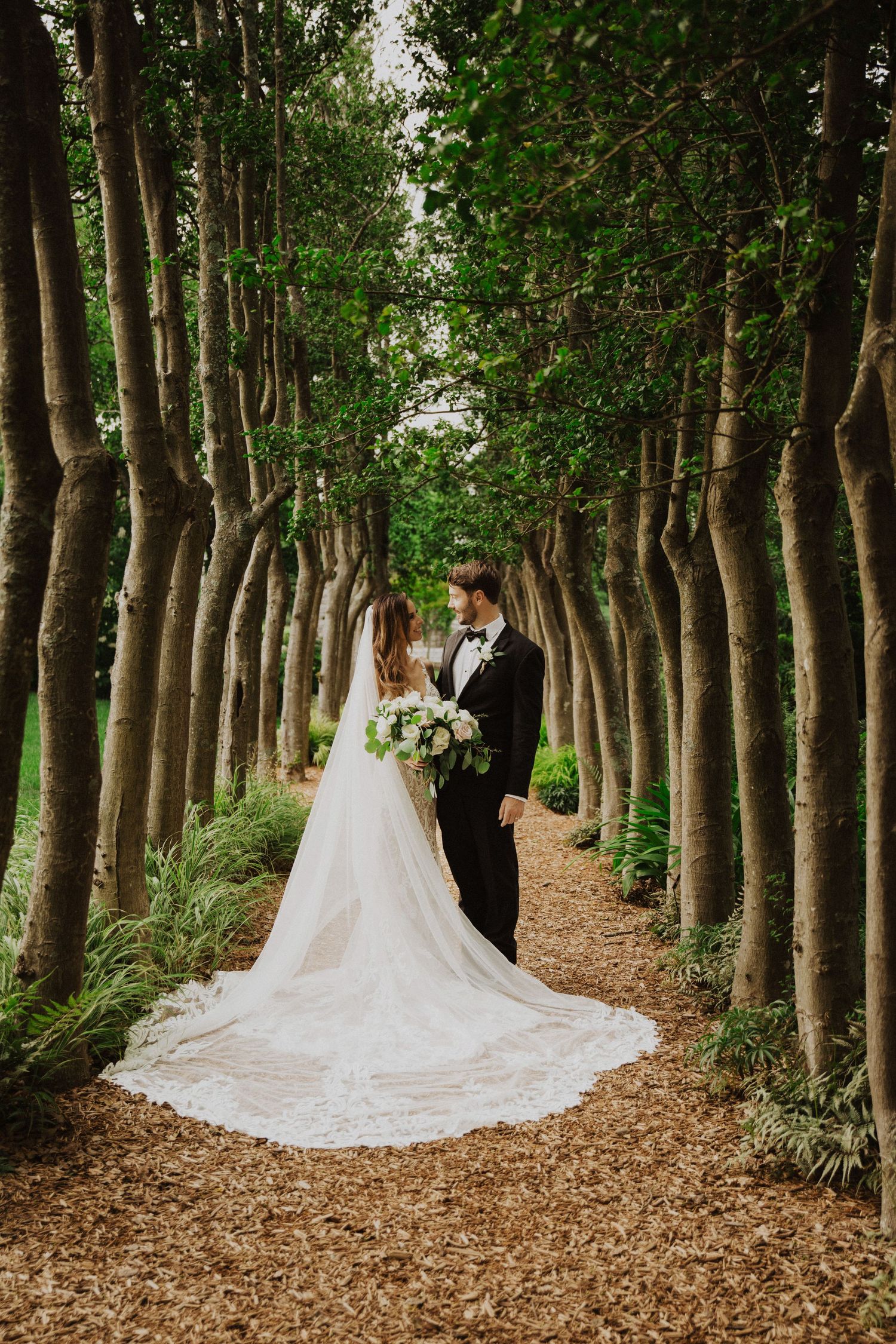 Wedding couple walking through tree-lined path with flowing white dress trailing behind.