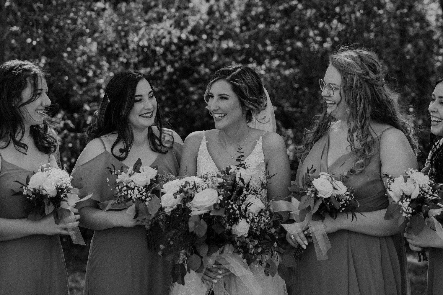 Bridesmaids in neutral dresses pose together holding white floral bouquets.