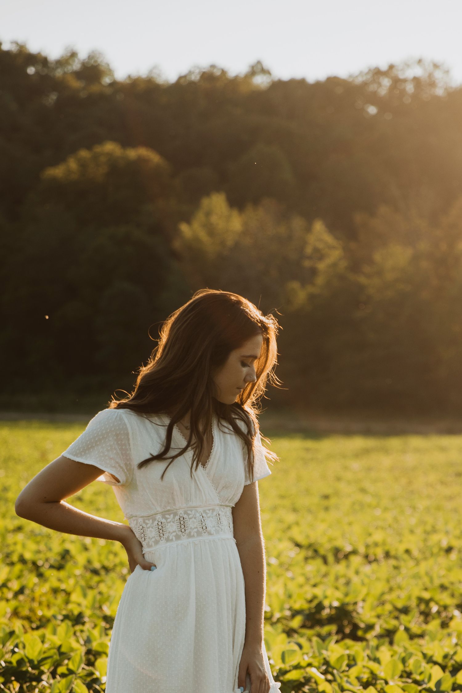 Young woman in white lace dress standing in sunlit green field with trees in background.