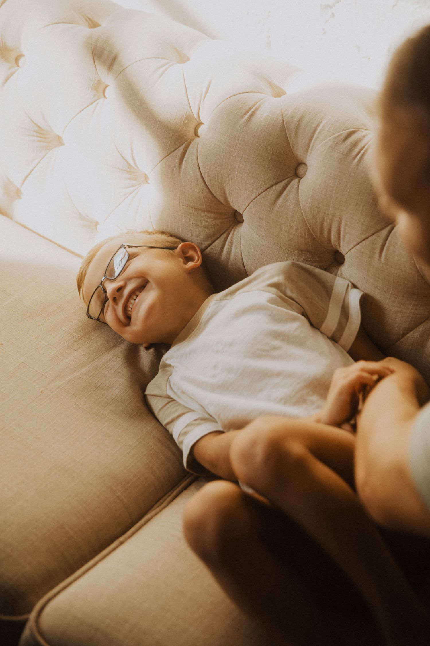 A smiling child wearing glasses lies on a beige tufted sofa in warm sunlight.