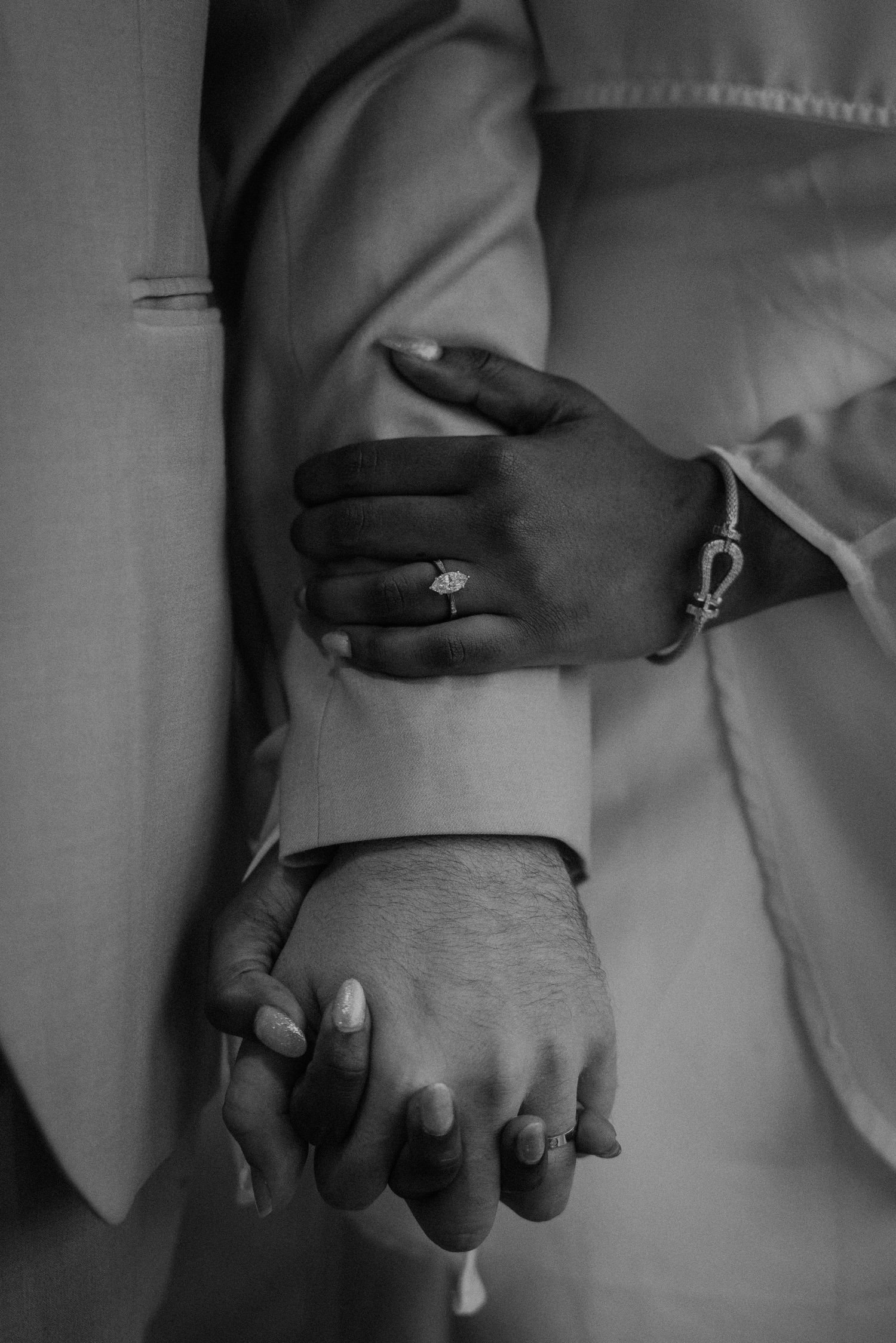 Black and white photo of an interracial couple holding hands, showcasing an engagement ring and bracelet.
