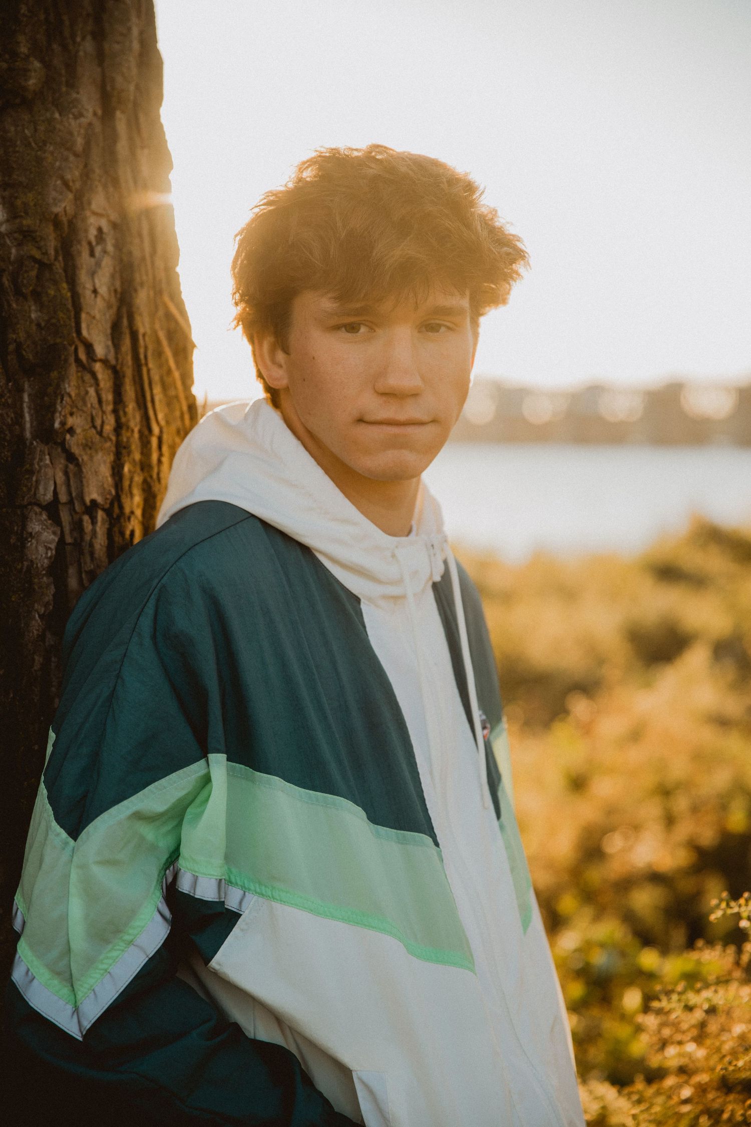 Teen boy in retro windbreaker jacket leans against tree at golden hour near waterfront.