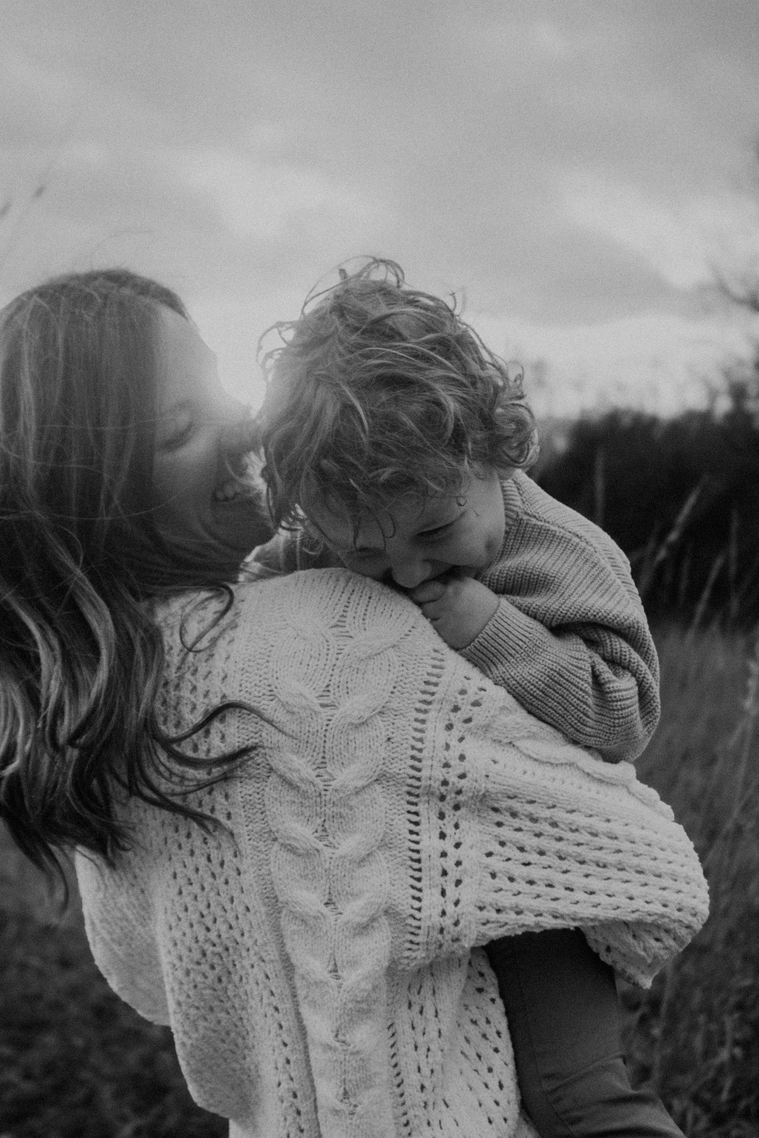 Black and white photo of a mother holding and kissing her curly-haired child outdoors in a field.