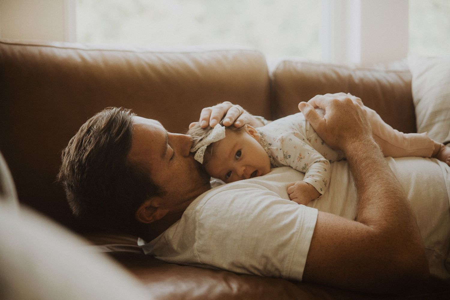 Father lying on brown leather sofa kissing newborn baby while cradling infant in his arms.