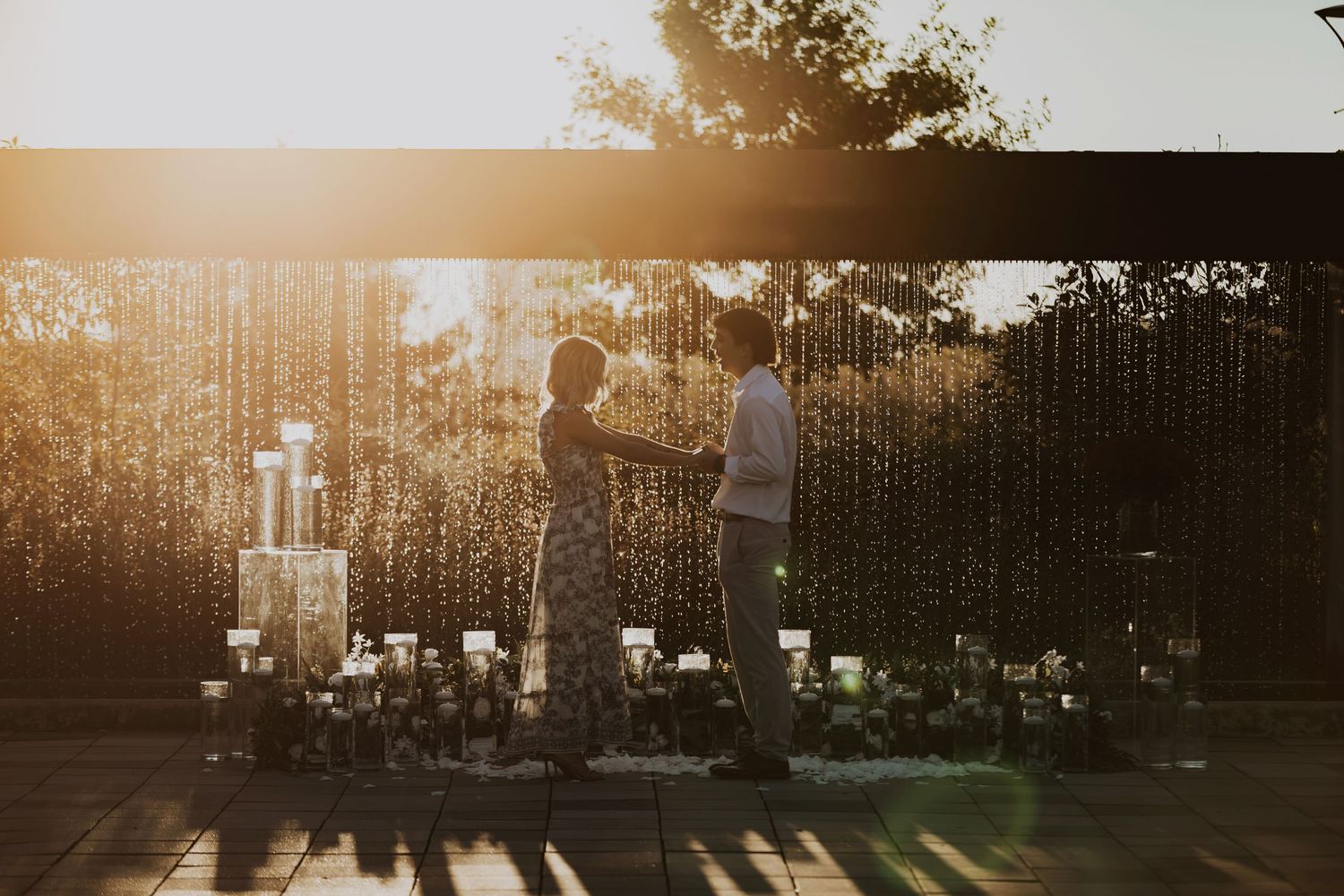 Couple silhouetted against golden sunset with string lights and candles creating romantic ambiance.