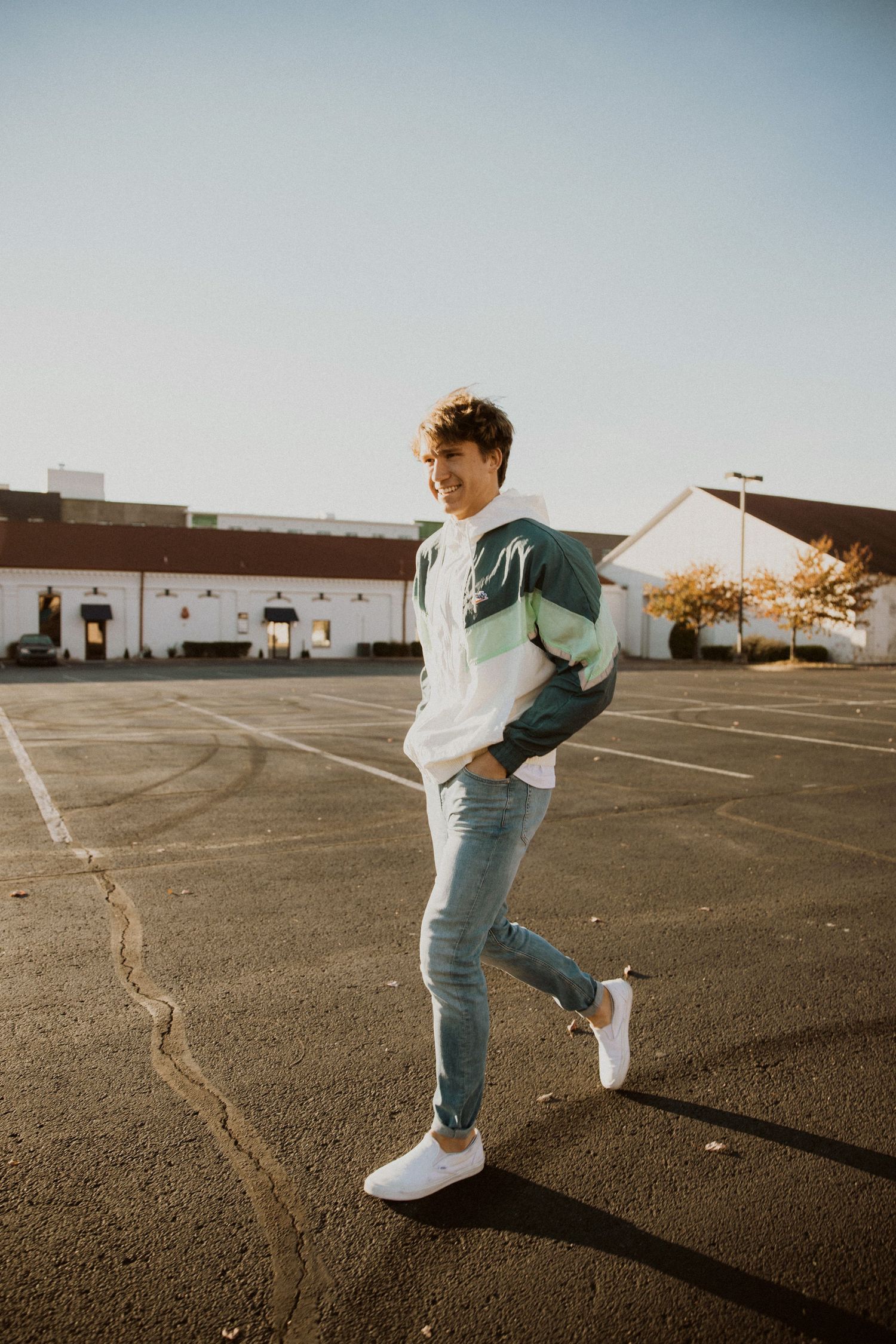 Young man in green colorblock sweatshirt and jeans poses confidently in sunny parking lot wearing white sneakers.