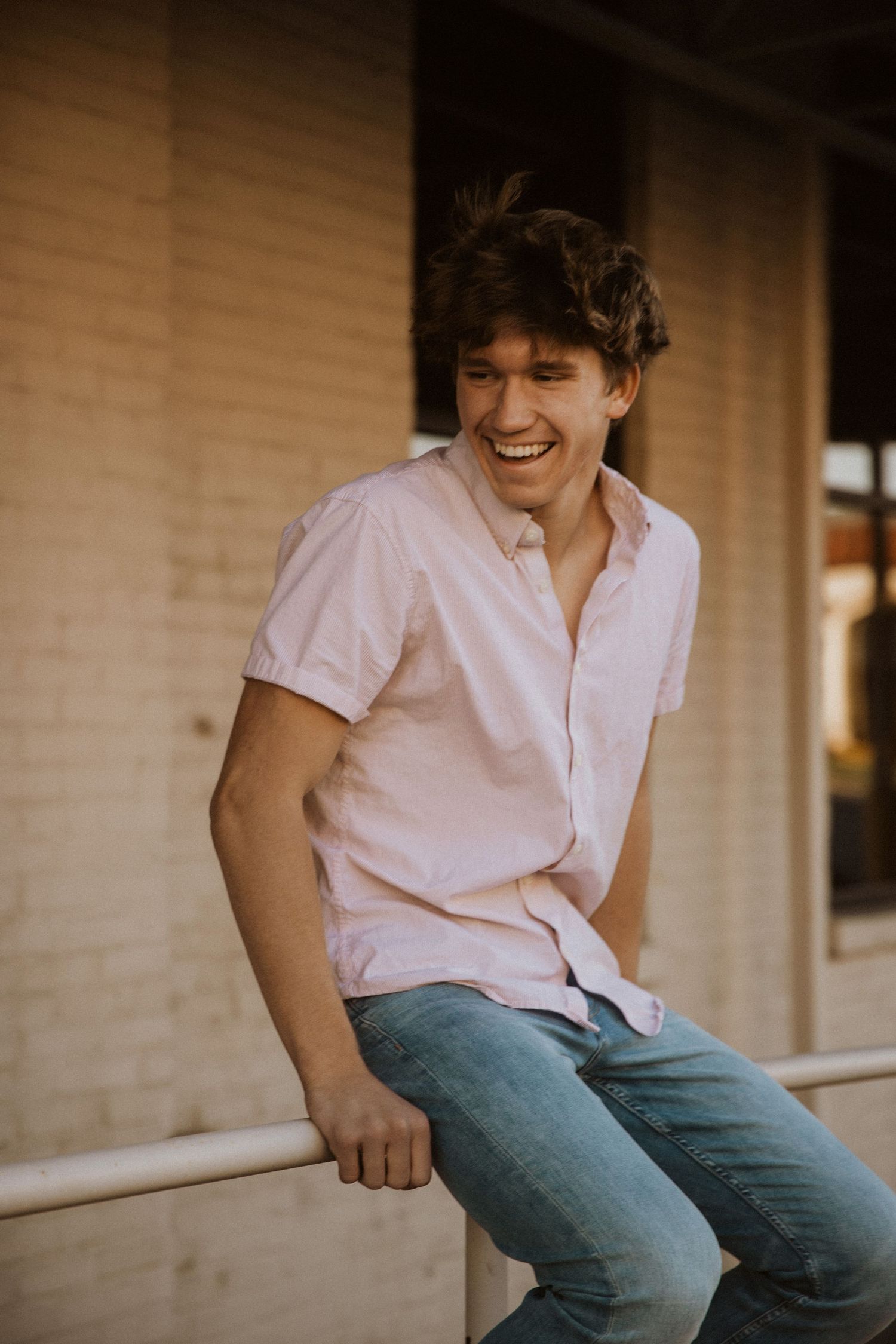 Young man in pink shirt and jeans laughing while sitting on a railing outdoors near a brick wall.