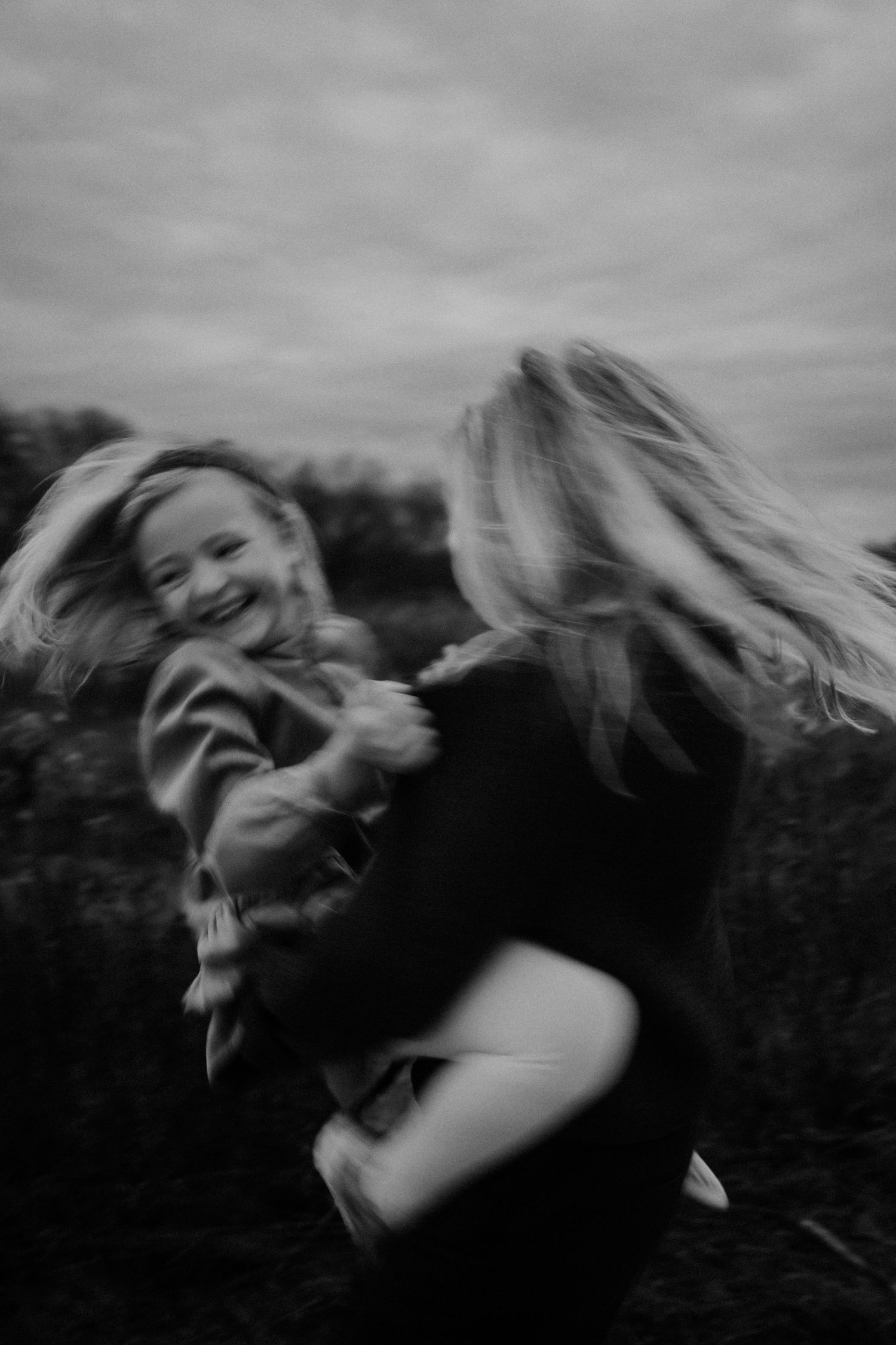Black and white photo of a laughing child being spun around by a woman outdoors on a cloudy day.