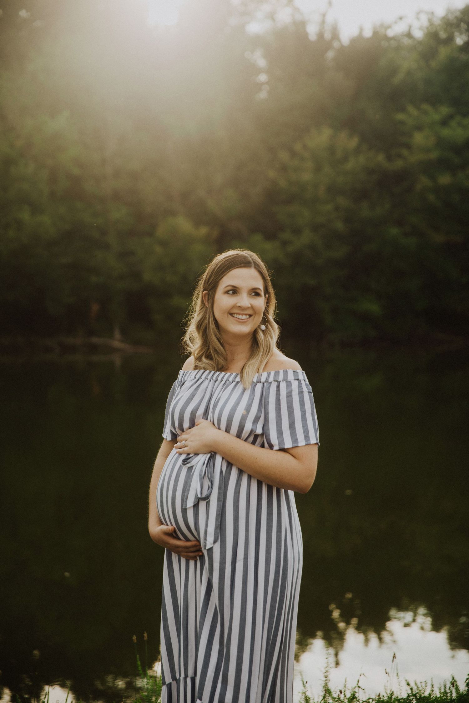Smiling pregnant woman in striped off-shoulder dress cradling baby bump by a sunlit pond surrounded by lush green trees.