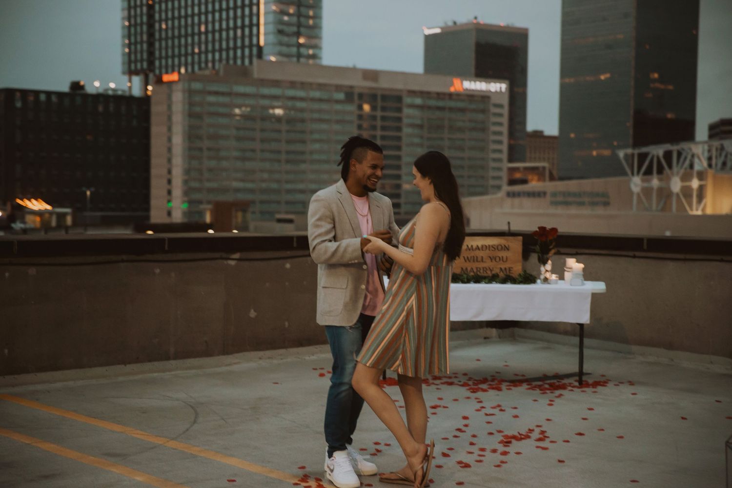 A romantic rooftop proposal setup with scattered rose petals and city buildings in background.