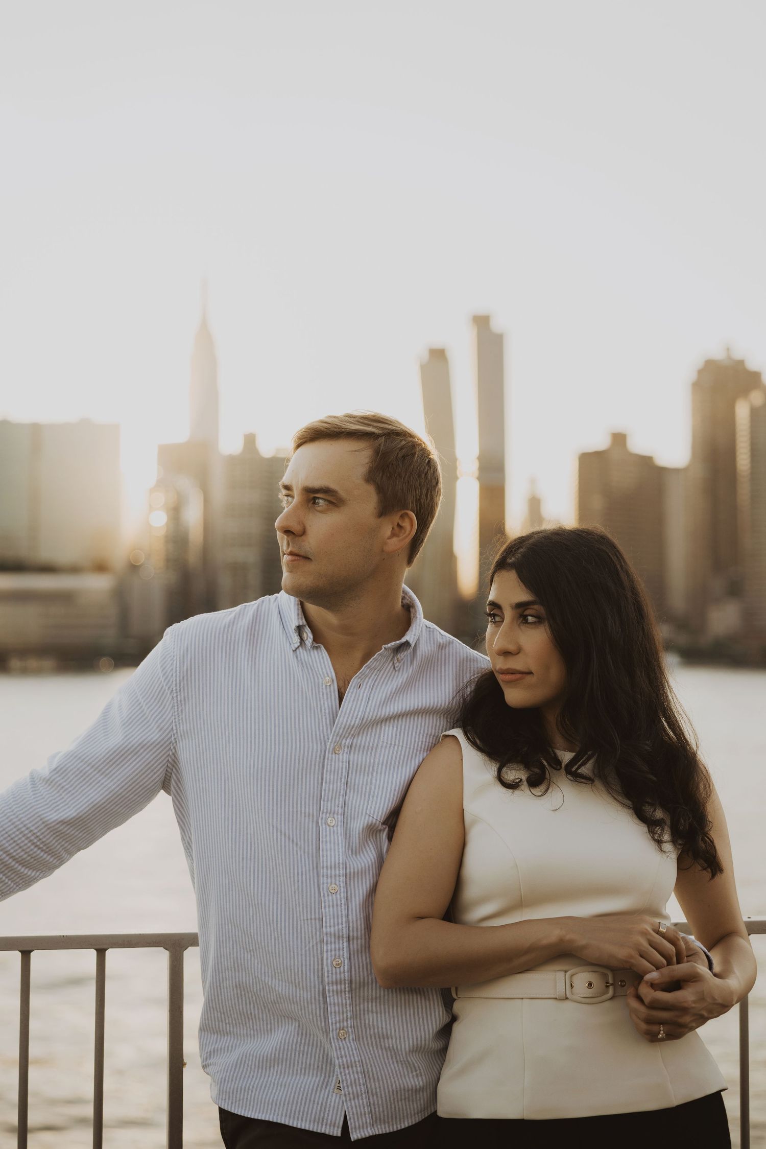 Couple embracing with NYC skyline and golden sunset backdrop, man in blue shirt, woman in white dress.