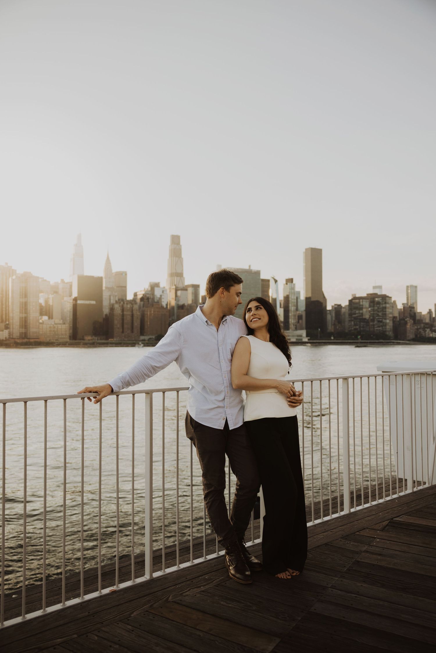 Couple embracing on a waterfront pier with the New York City skyline and East River at golden hour.