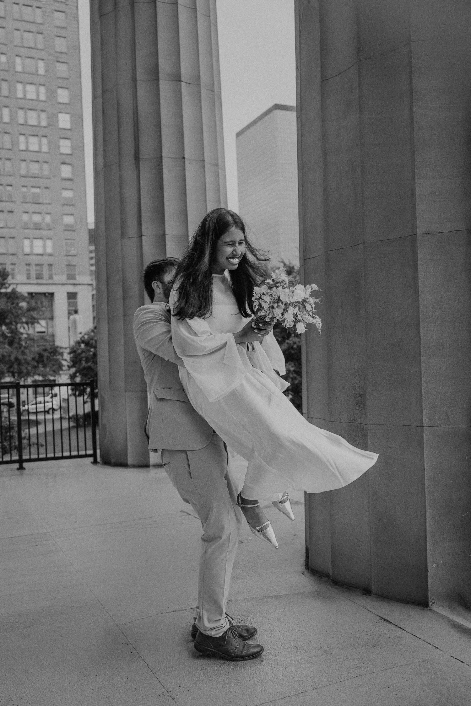 Black and white photo of groom lifting bride in white dress holding bouquet on urban building steps.